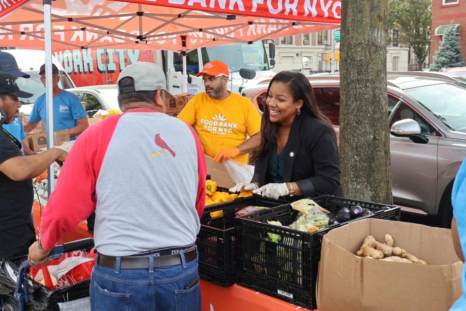 People at a food distribution event with a woman smiling and handing vegetables to a man, volunteers working behind the table, and a red canopy with 'Food Bank for NYC' written on it.