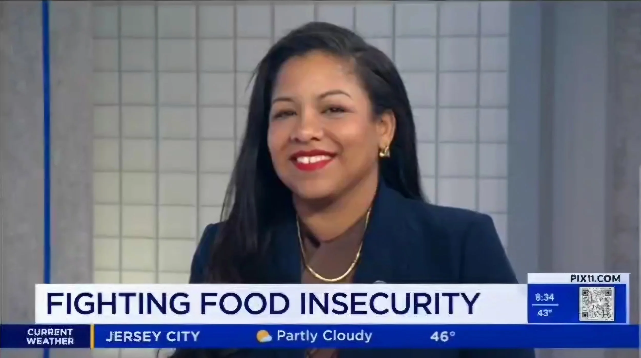 A woman with long dark hair, wearing a black blazer, jewelry, and red lipstick, smiles during a news interview about fighting food insecurity. The news banner indicates it is from Jersey City and the time is 8:34.