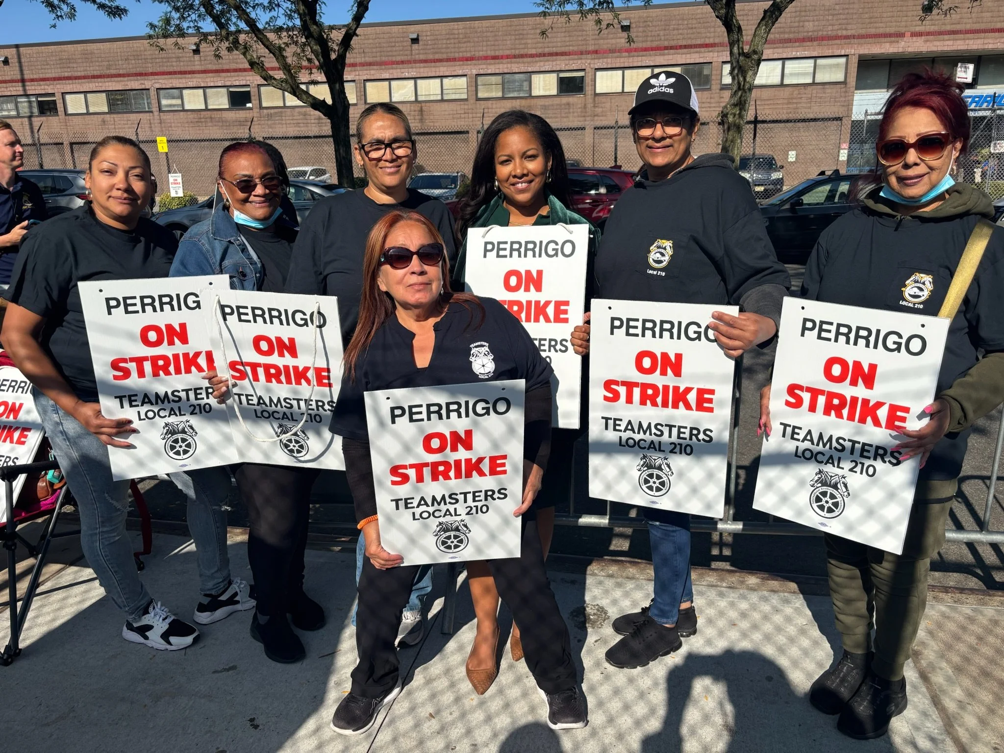 Group of women holding protest signs that read 'PERRIGO ON STRIKE' and 'TEAMSTERS LOCAL 210' outside, with some women wearing black clothing and sunglasses, in an outdoor parking lot area.