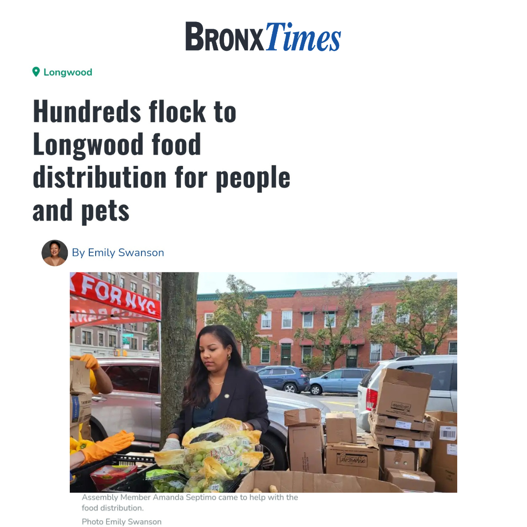 A woman with dark hair wearing a black blazer is standing outside near parked cars and trees, surrounded by cardboard boxes and food supplies, participating in a food distribution event.