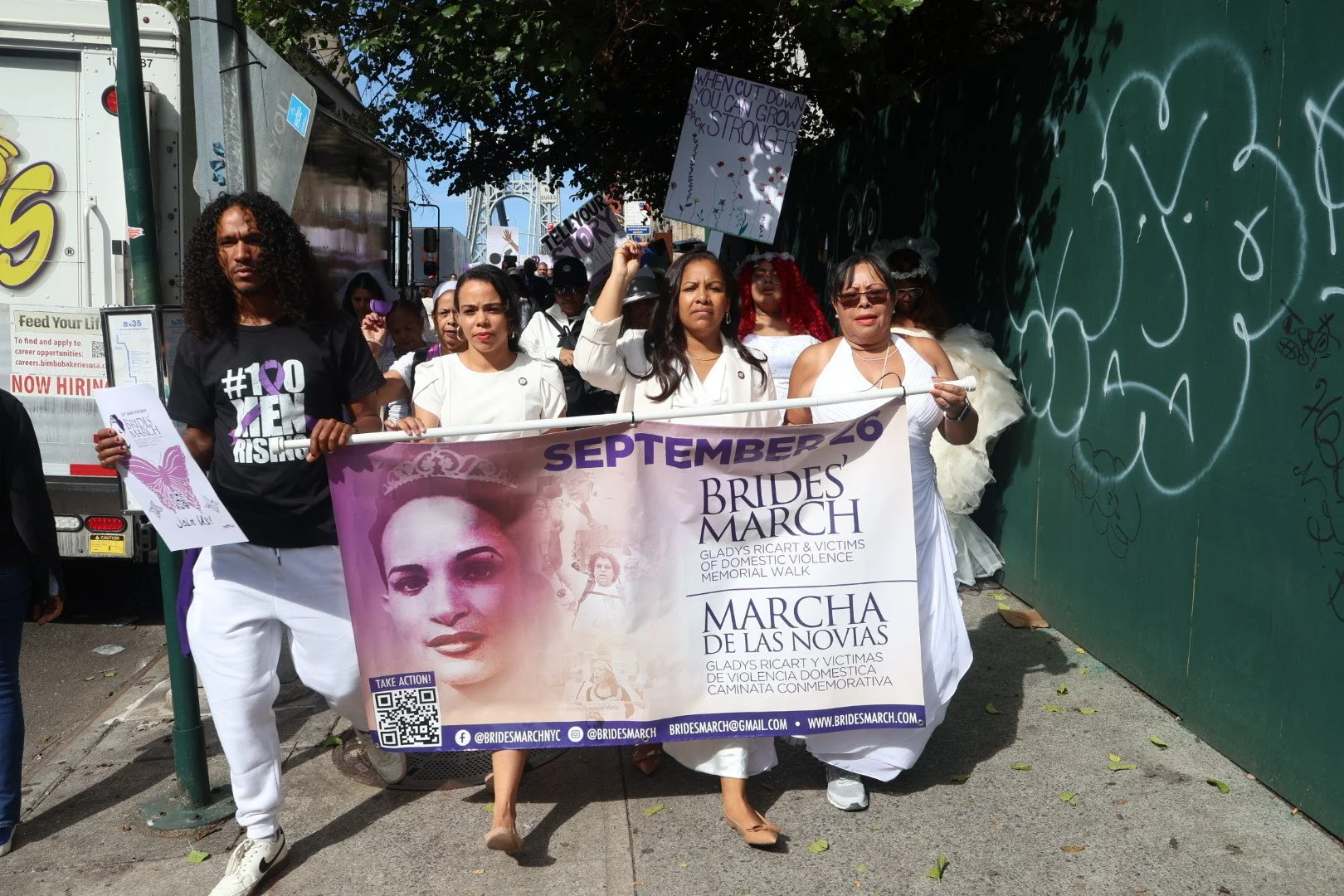 Women participating in the Brides March parade, carrying a banner with a photo of Gladys Ricart, against domestic violence, walking along a city sidewalk with others and graffiti-covered wall nearby.