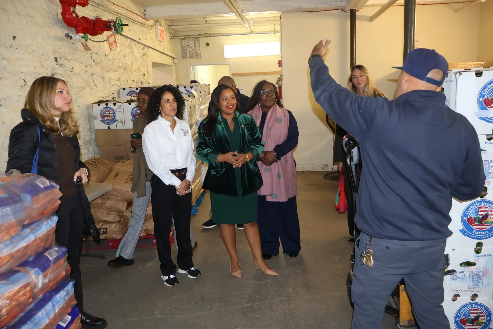 Group of women listening to a man in a warehouse with stacked boxes and supplies.