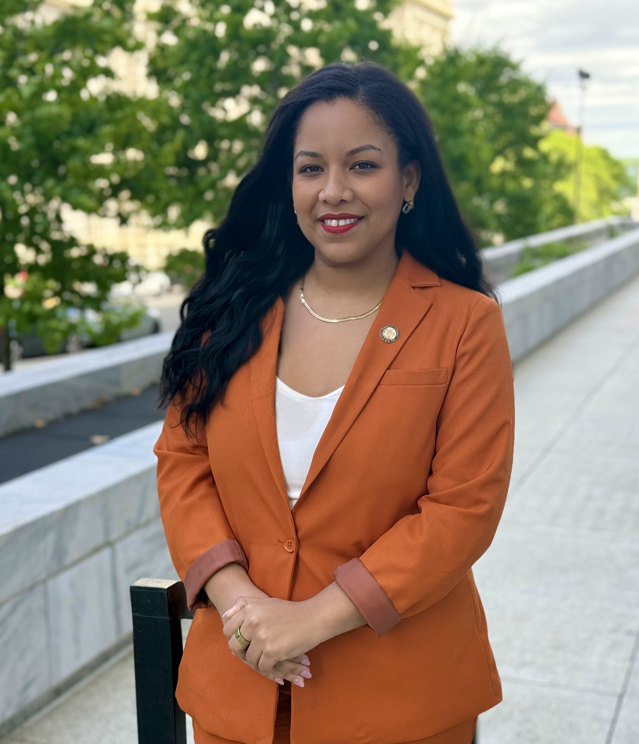 A woman with long black hair and a bright smile standing outdoors, wearing an orange blazer over a white shirt, with a pin on her lapel, on a walkway with green trees in the background.