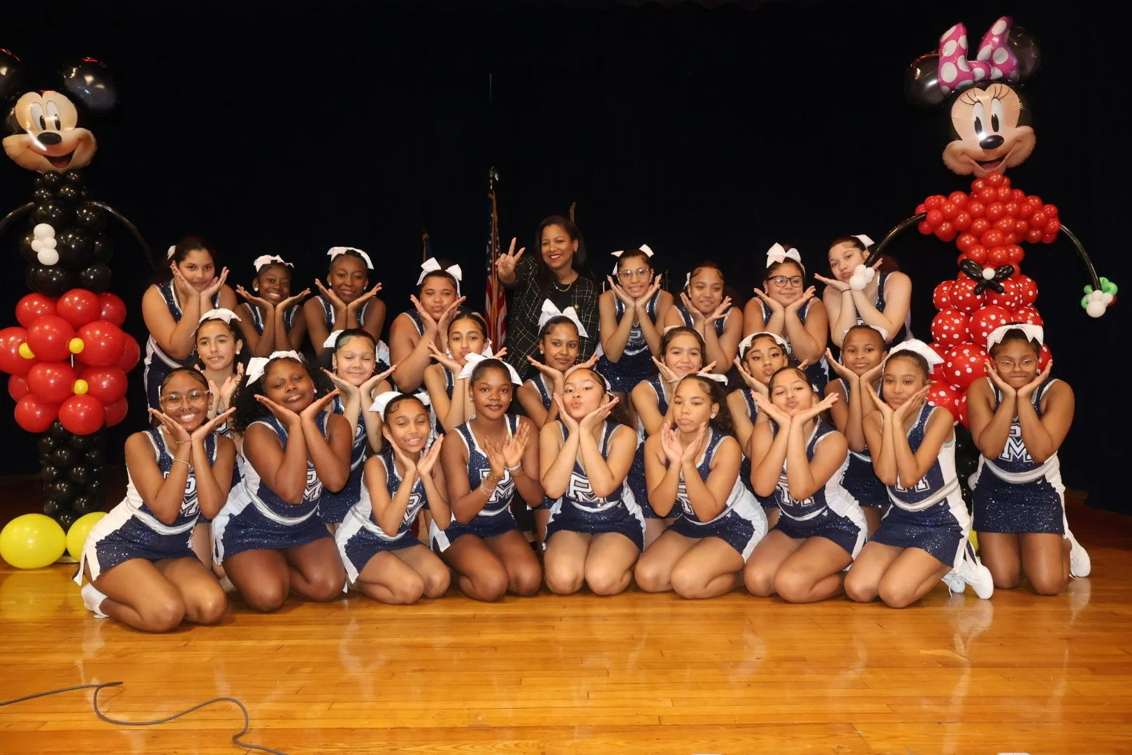 Group of young cheerleaders in navy and white uniforms posing with cheer coach, surrounded by large Minnie Mouse balloon figures on a stage with a black curtain background.