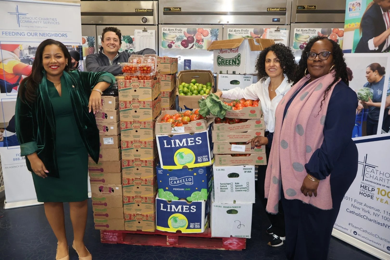 Four women and one man standing around a large stack of boxes of fruits and vegetables, including tomatoes, limes, and greens, at a charity event.