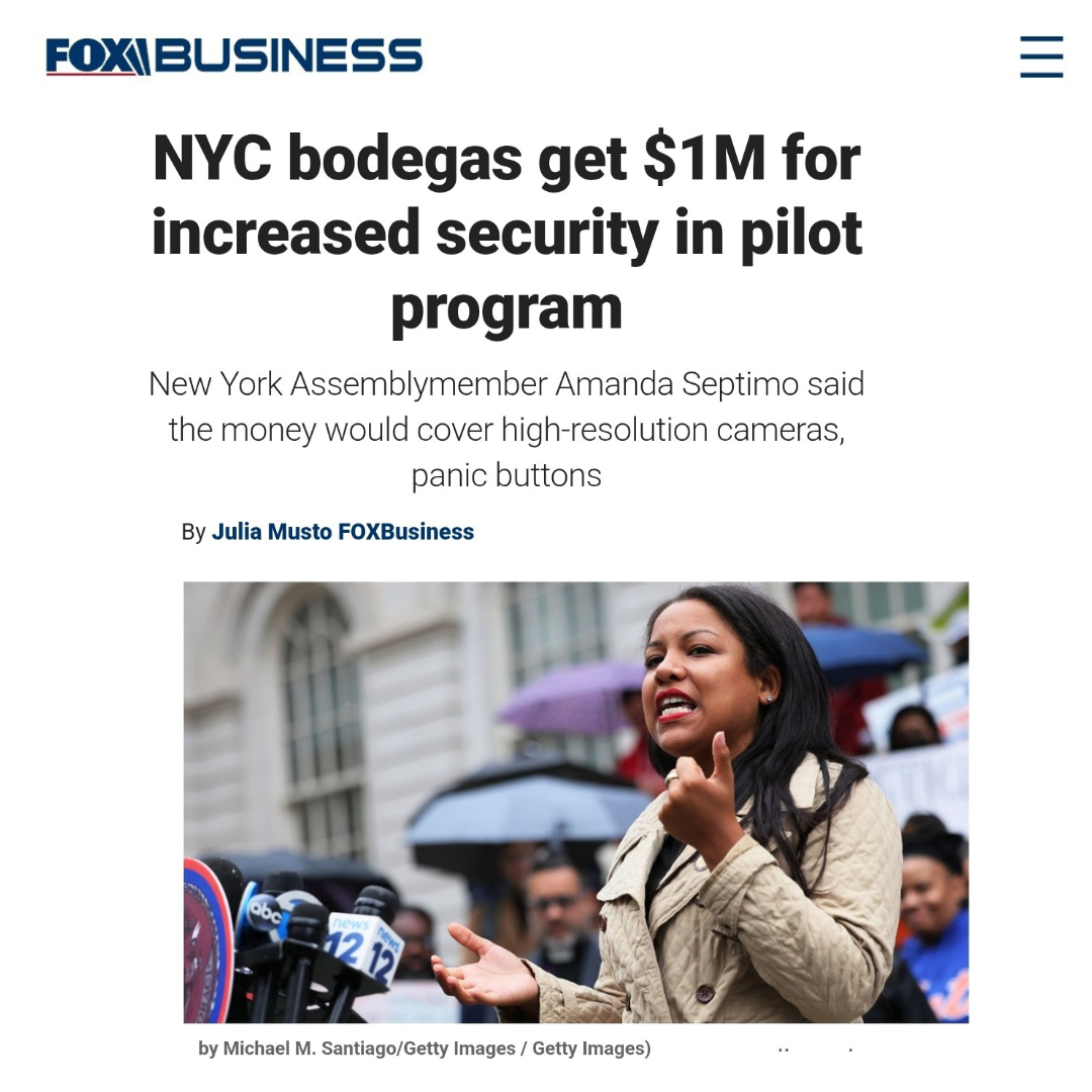 A woman speaking at a press conference outdoors during daytime, with microphones and reporters in front, and people holding umbrellas in the background.