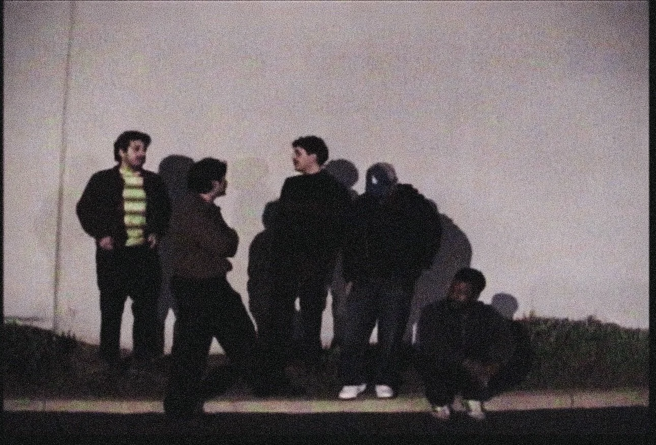 Six young men standing and sitting in a dimly lit room, some facing each other and others looking away, with a blank white wall in the background.