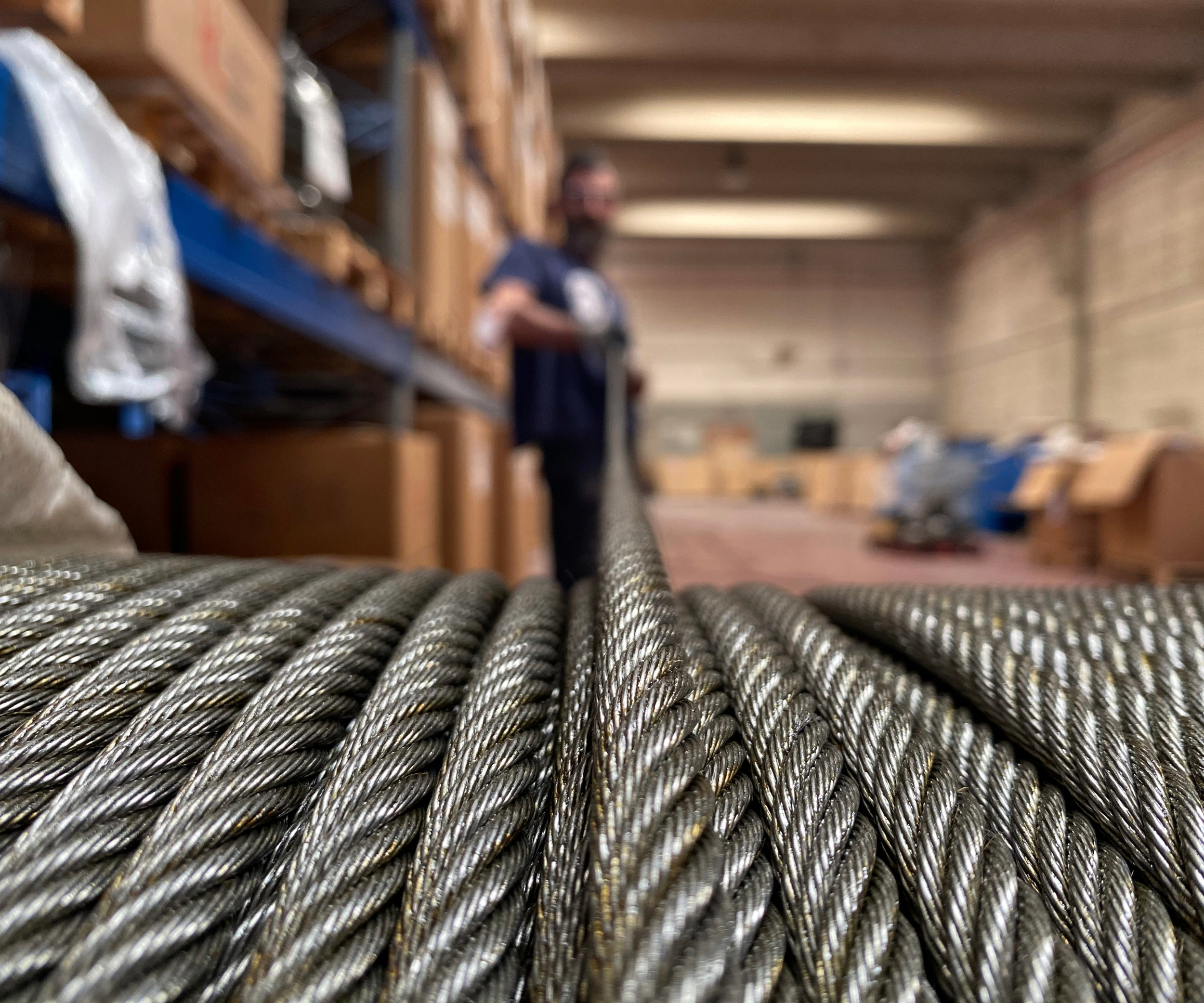 Close-up of a steel wire rope with a worker blurred in the background in a warehouse setting.
