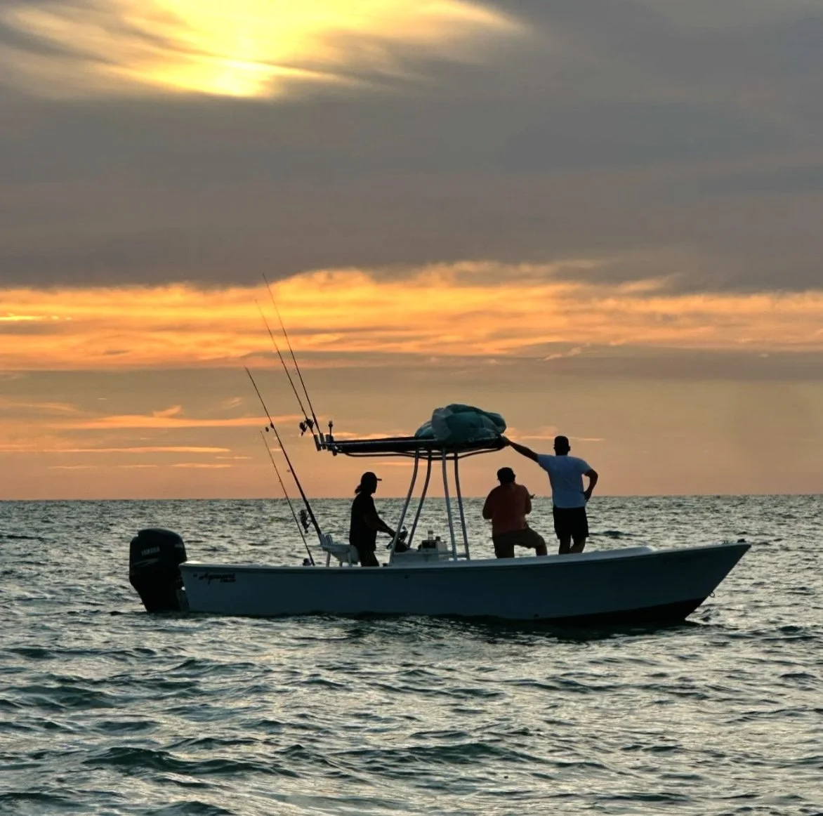 Silhouettes of three people fishing on a boat during sunset on the ocean.