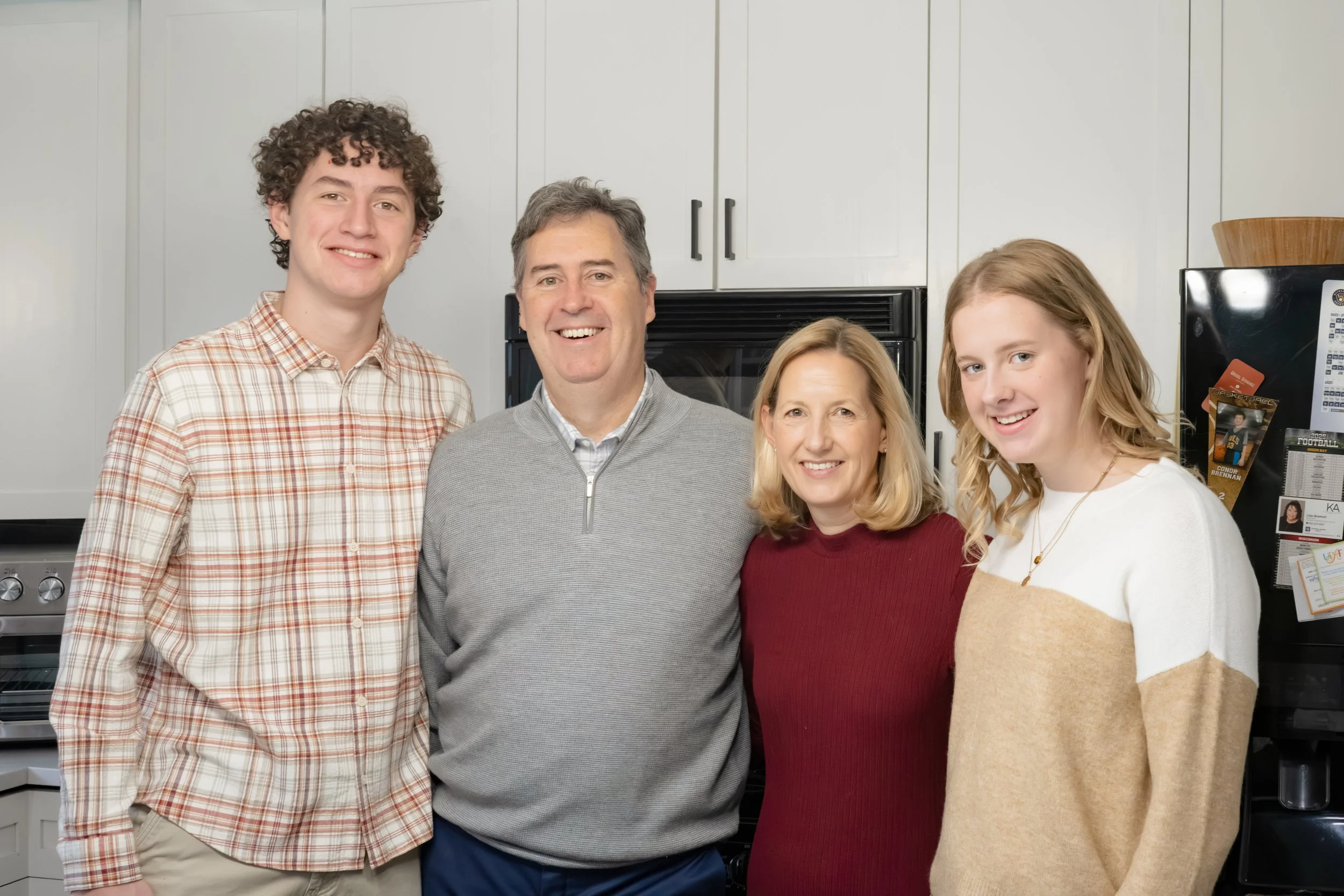 A family of four standing together in a kitchen, smiling at the camera. The family includes two young adults and two older adults, with a white kitchen cabinet and appliances in the background.