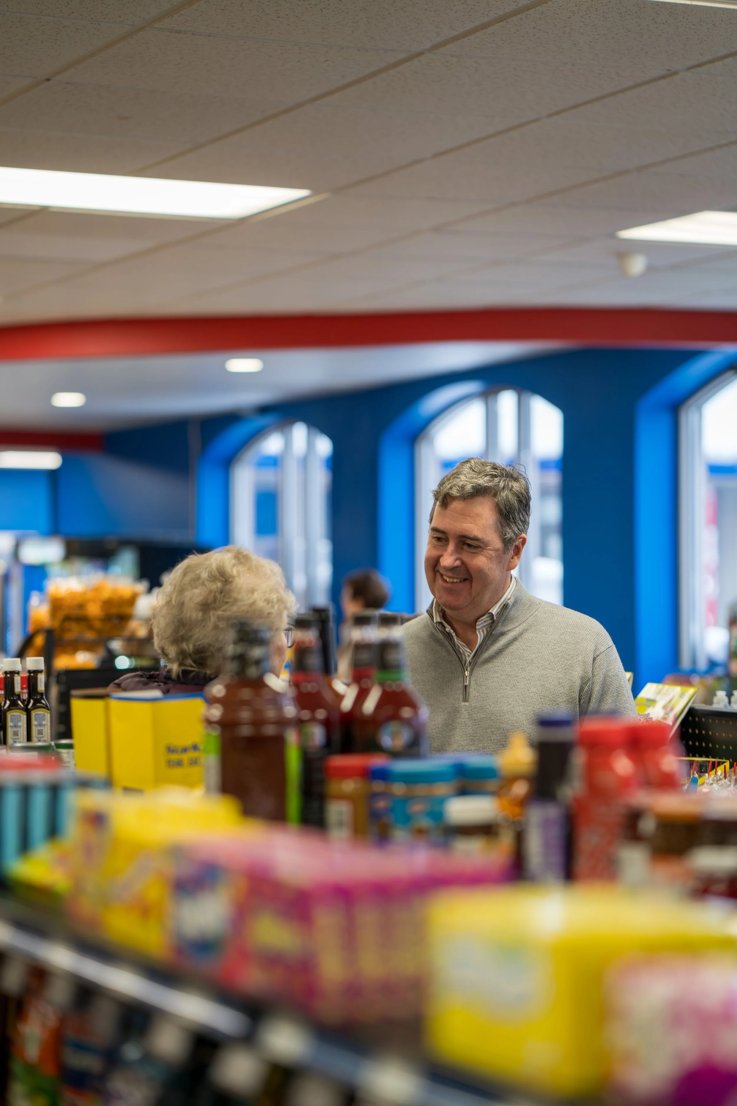 A man shopping in a grocery store, smiling and talking to an older woman, with shelves of food products in the foreground.