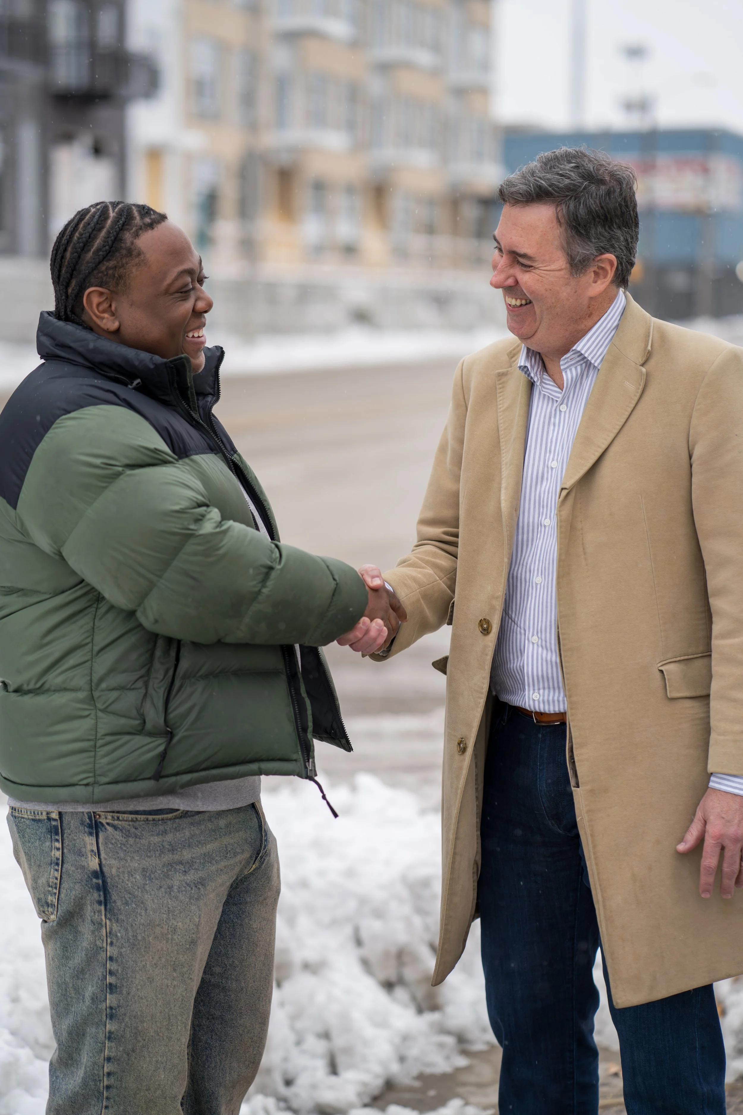 Two men shaking hands outdoors in a snowy, urban environment, smiling at each other.