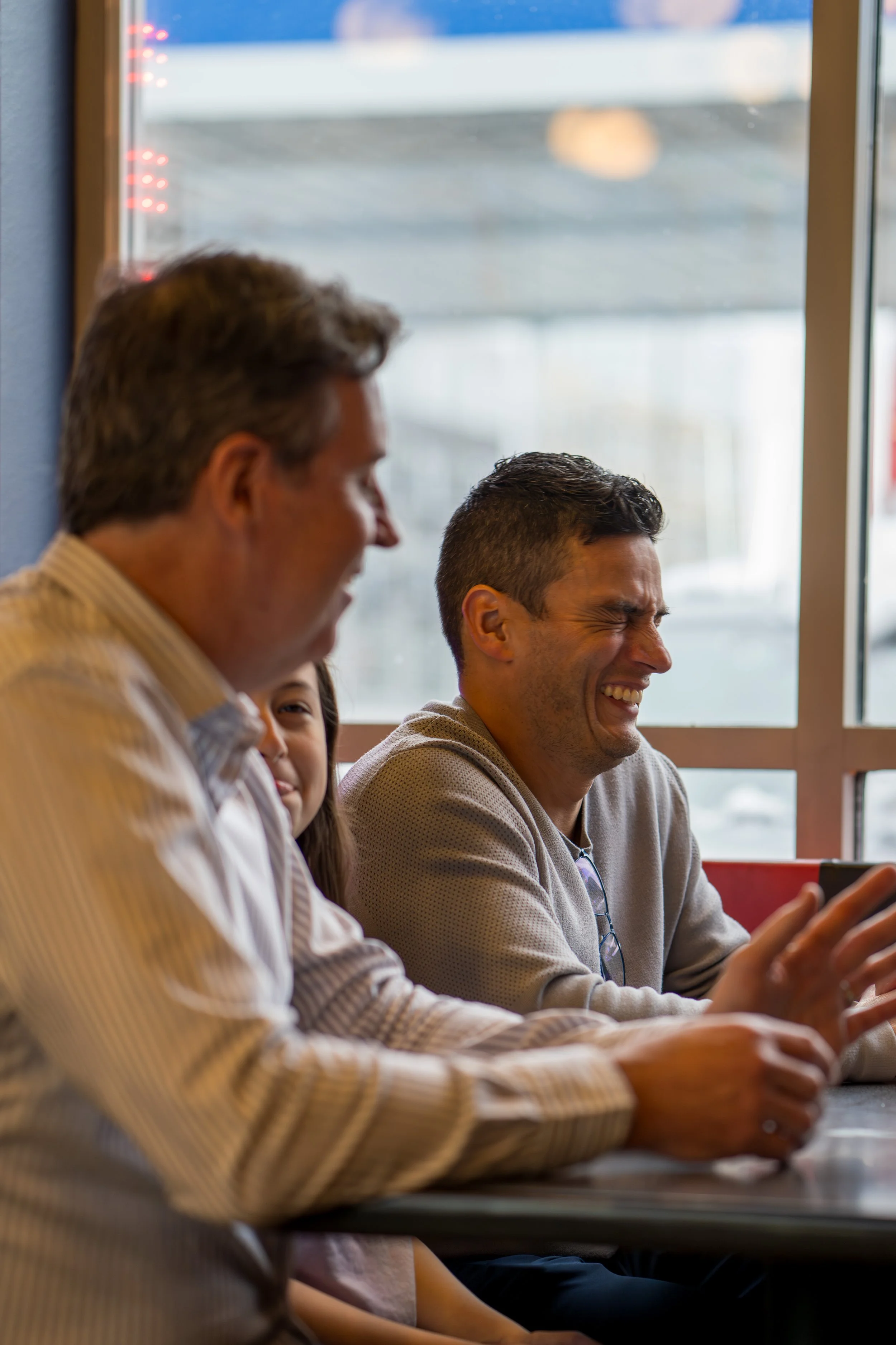 Three people sitting at a table inside a restaurant, laughing and smiling, with a large window behind them showing blurred outdoor scenery.