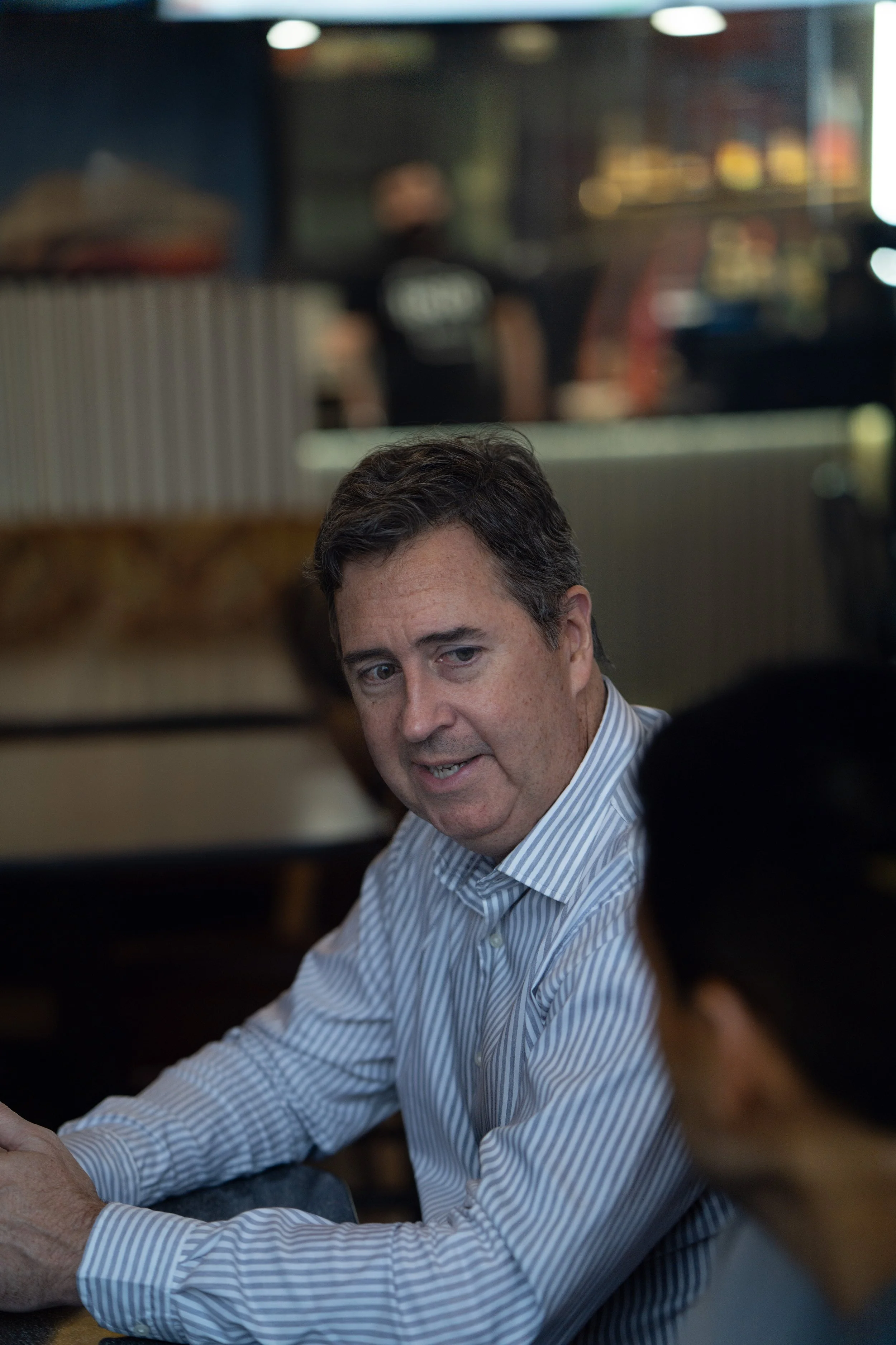 A man with brown hair wearing a light striped shirt is sitting at a table inside a restaurant or cafe, engaged in conversation with another person who is partially visible in the foreground. The background features a blurred kitchen or service area w