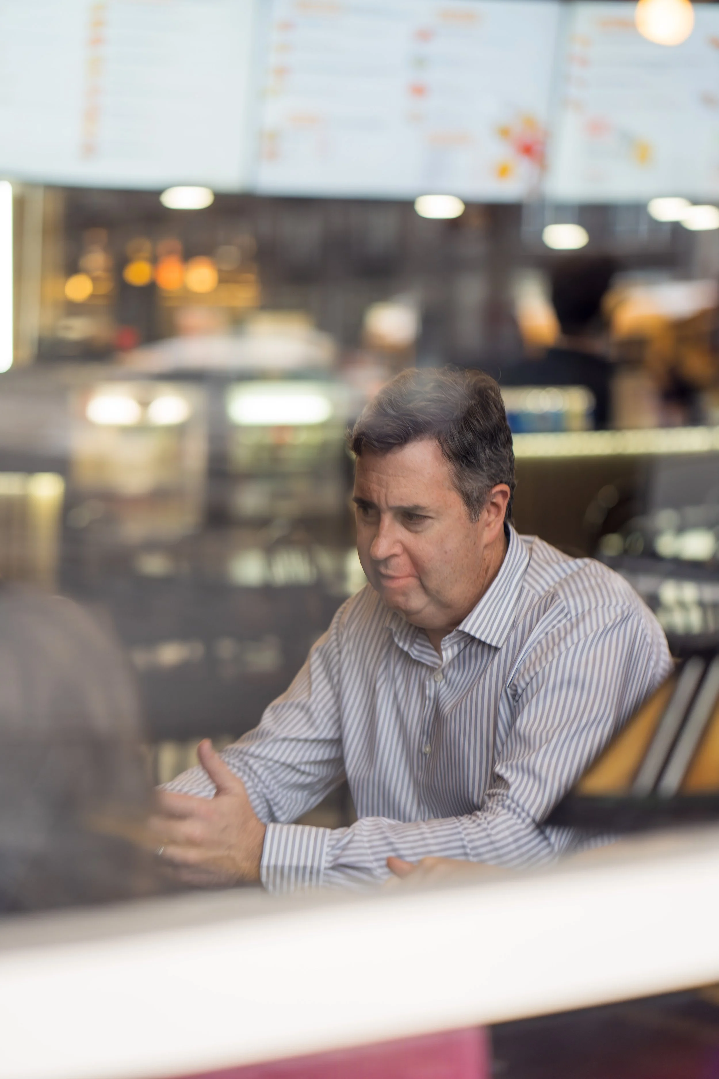 A man in a striped collared shirt sitting inside a café or restaurant, looking down with a thoughtful expression, with a blurred menu board and interior lights in the background.