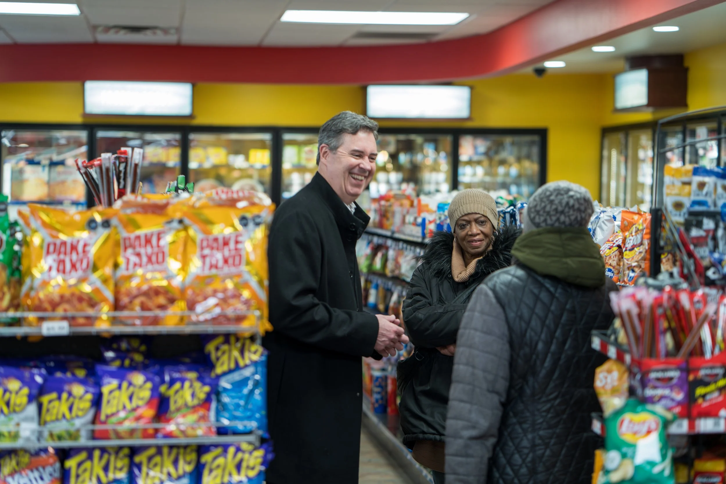 Three people in a grocery store aisle, two women and a man, all dressed warmly, engaged in conversation and smiling. The store has bright yellow walls and snack shelves.