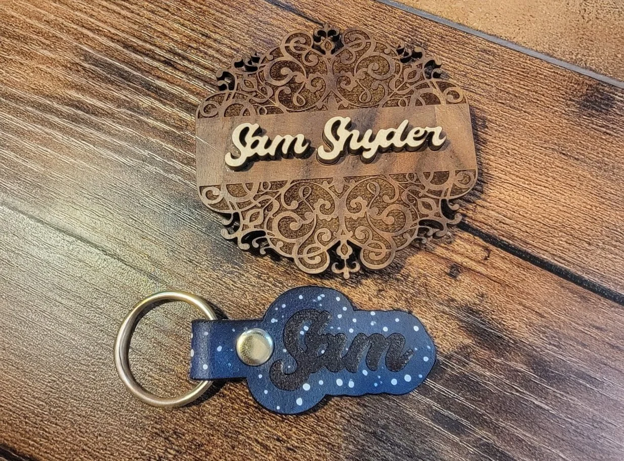 A decorative wooden nameplate with intricate cutout designs reading 'Sam Snyder' and a matching keychain with the same name on a wooden surface.