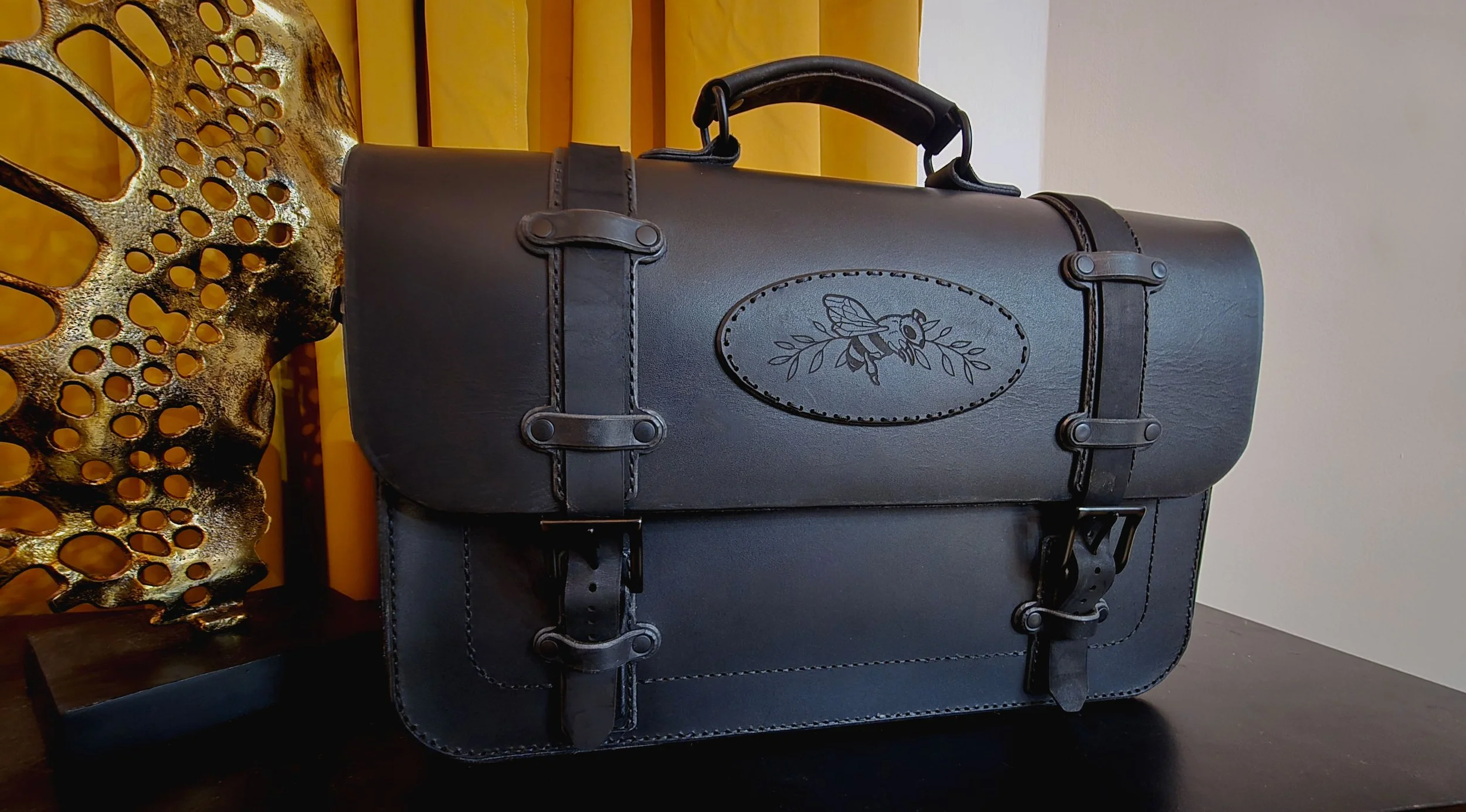 Black leather briefcase with decorative stitching and a bee and leaf design on the front.