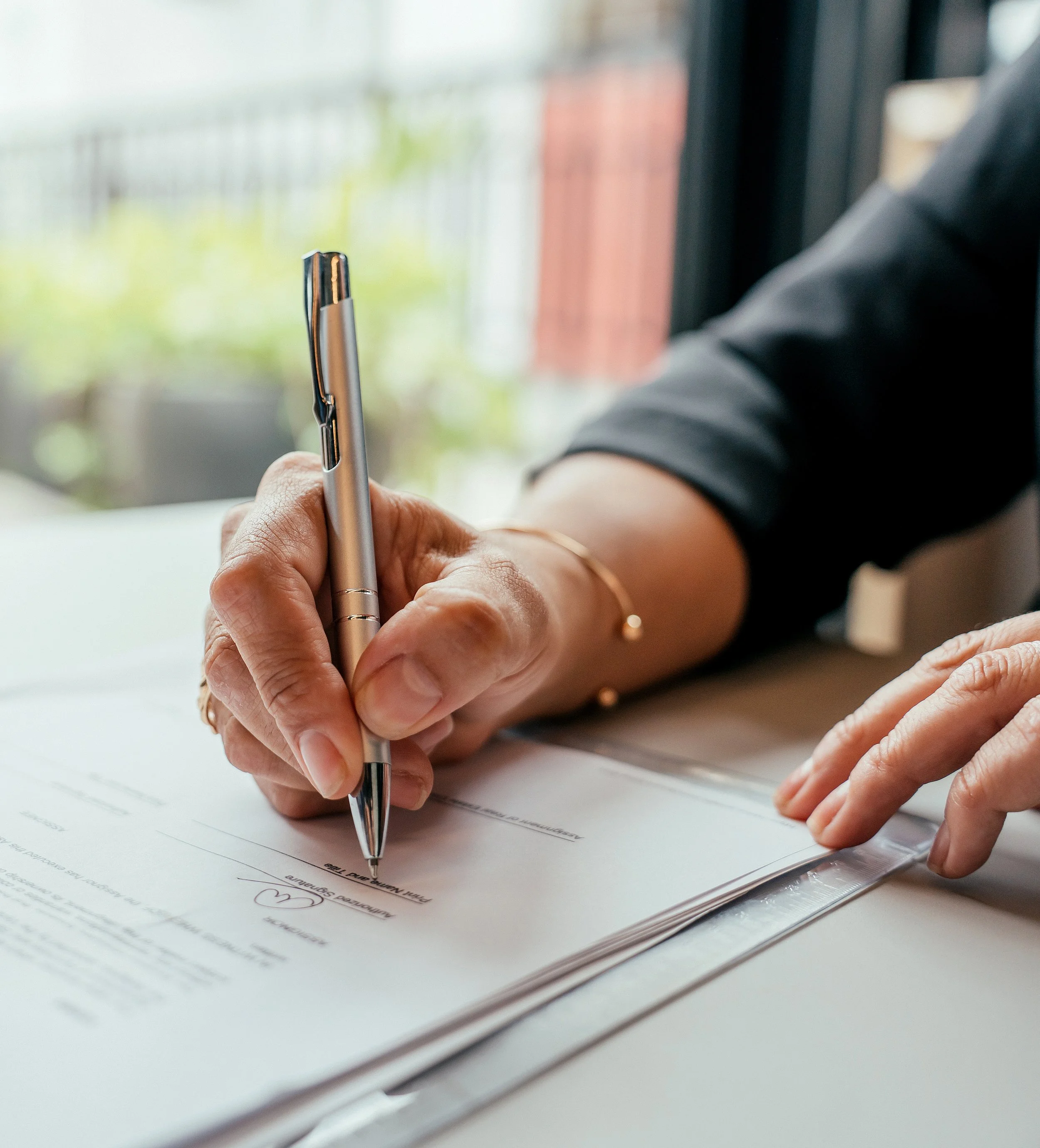 Person signing a document at a desk with a pen, near a window.
