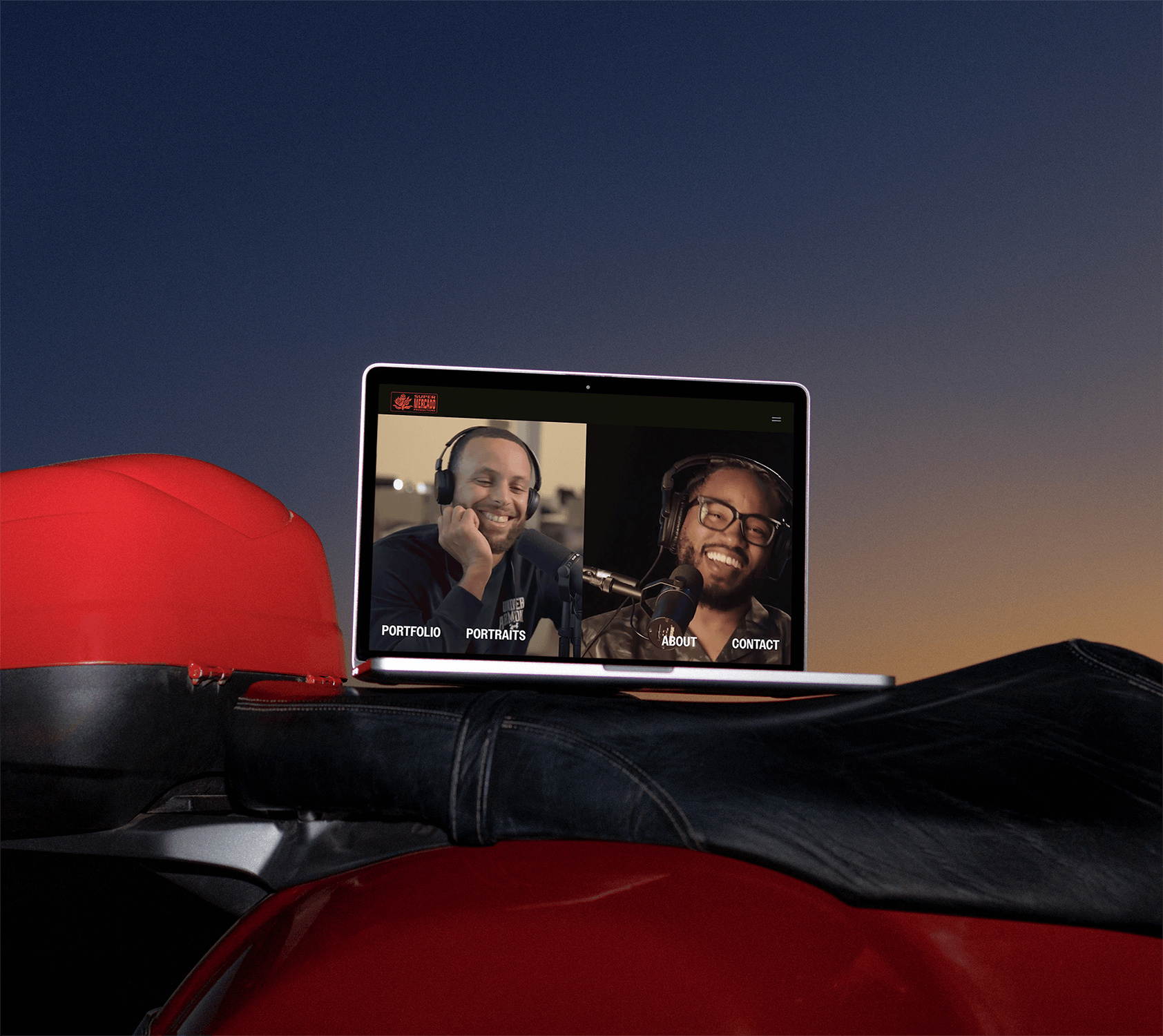 Laptop displaying a podcast with two men with headphones, smiling and speaking into microphones, on a red motorcycle tank with a sunset or dusk sky in the background.