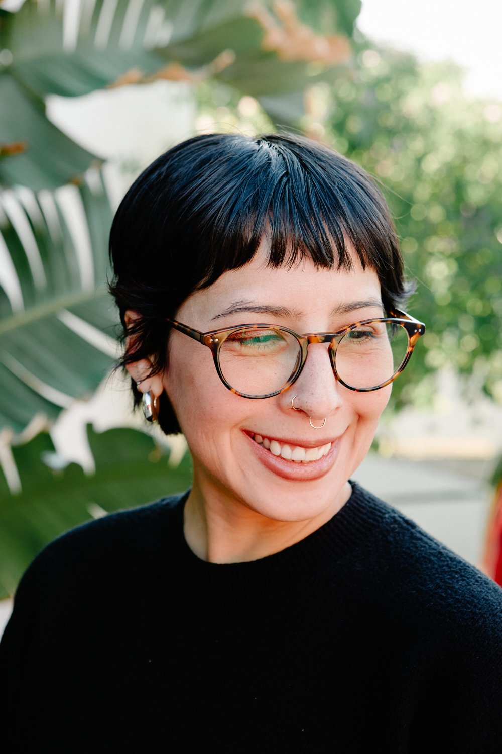 Smiling person with short black hair, tortoiseshell glasses, and a nose ring, standing outdoors near green foliage.