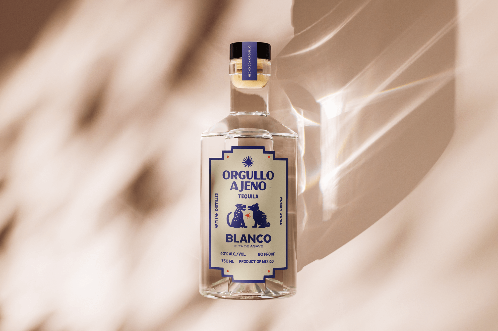 A clear glass bottle of tequila labeled 'Orgullo A Jeno Blanco' with artwork of two stylized animals and information about alcohol content, origin, and volume, placed against a soft, neutral background with shadows.