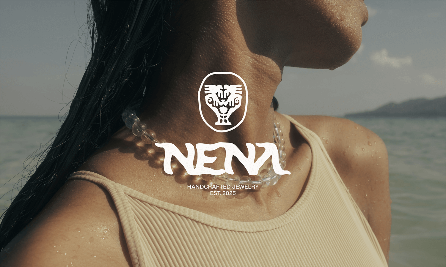 Close-up of a woman wearing a beige tank top and a beaded necklace at the beach, with water and sky in the background, and a logo with a lion face and the word 'NENA' overlayed on the image.