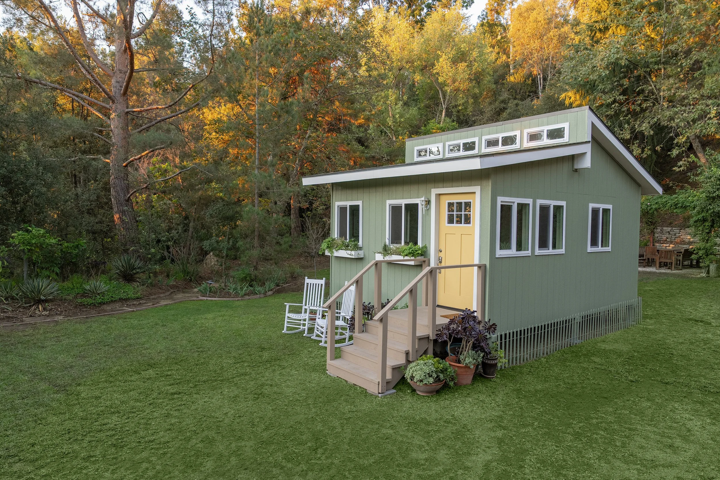 A small green house with a yellow door and multiple windows is situated in a lush yard surrounded by trees with autumn foliage. There are white rocking chairs and potted plants near the entrance, with a forested background.