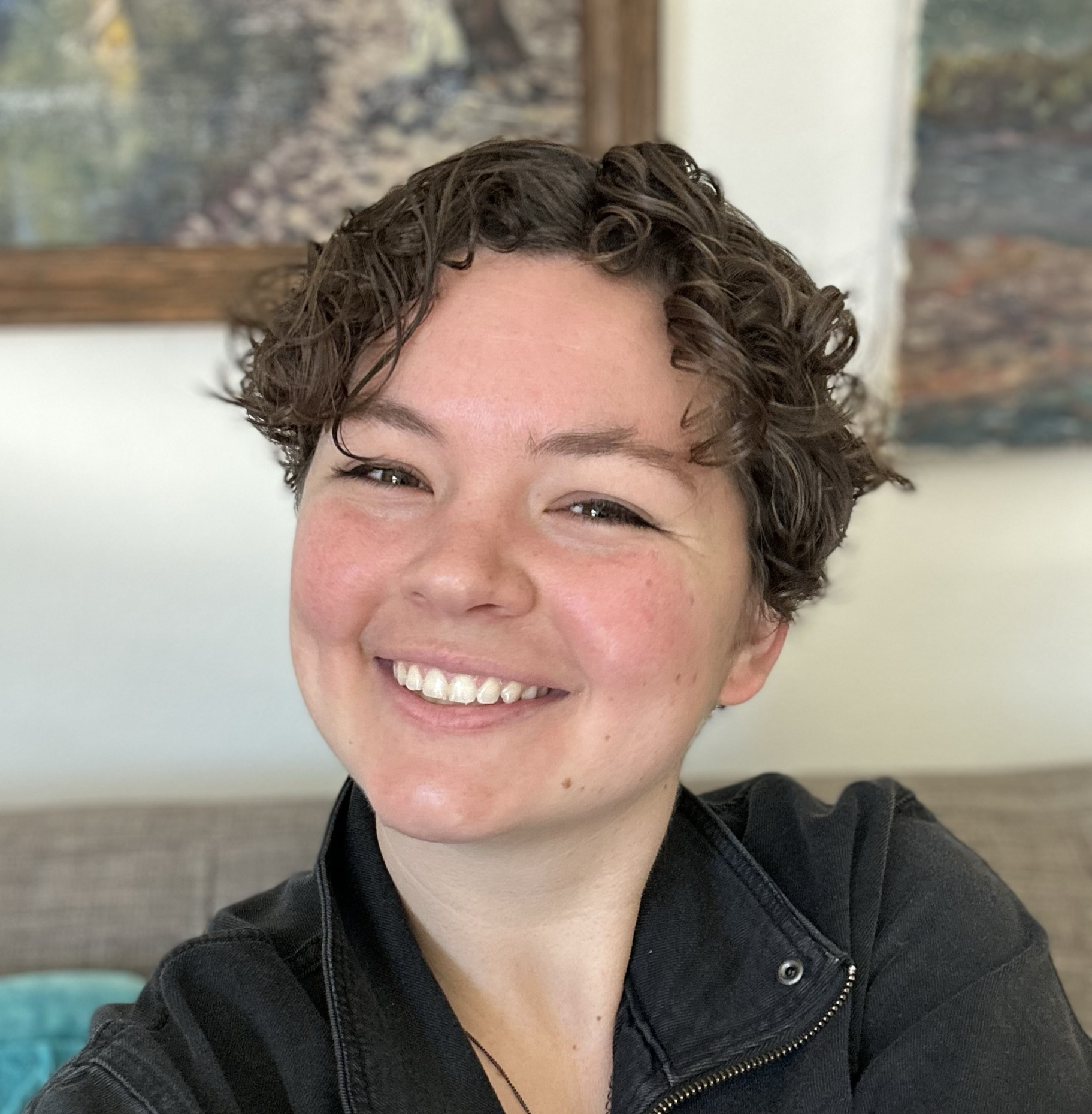 A woman with short, curly brown hair smiling and looking at the camera, wearing a black jacket, with a blurred background of art on the wall and a window.