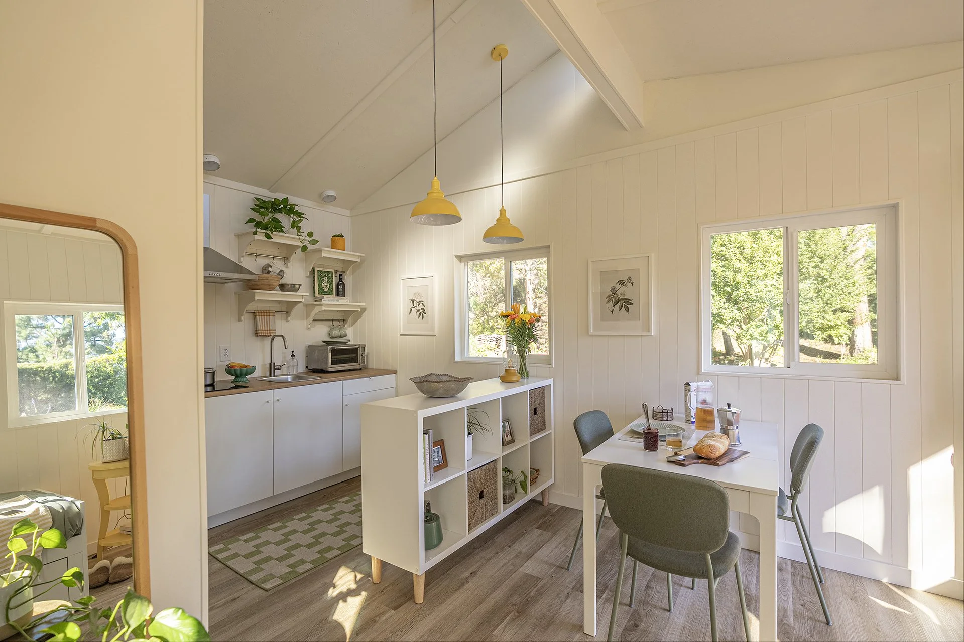 Bright kitchen and dining area with white walls, two yellow pendant lights, dining table with four chairs, windows showing greenery outside, and kitchen with open shelves, microwave, and sink.