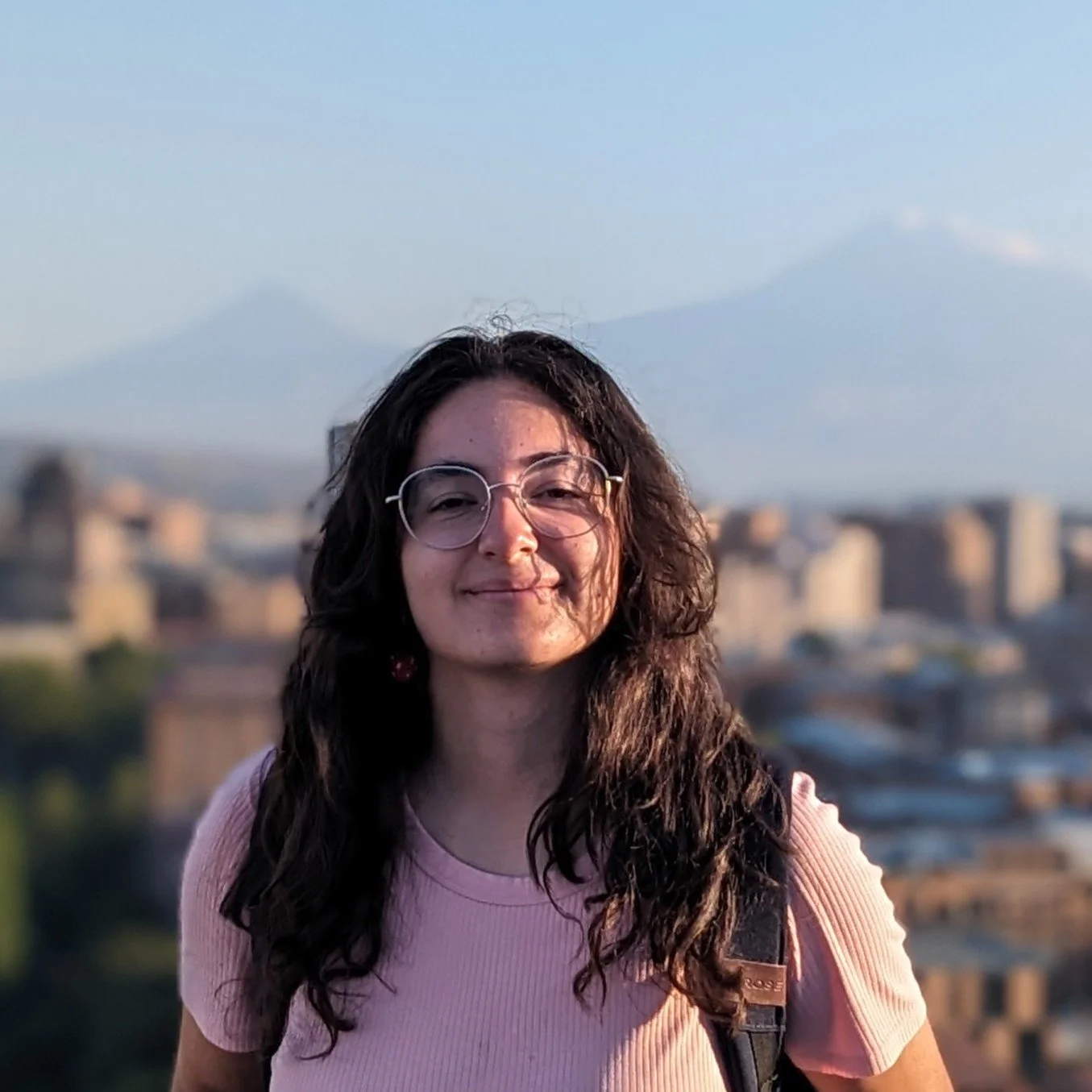 A woman with dark curly hair, glasses, and earrings is smiling, wearing a pink top and carrying a backpack, with an urban cityscape and mountains in the background during sunset.