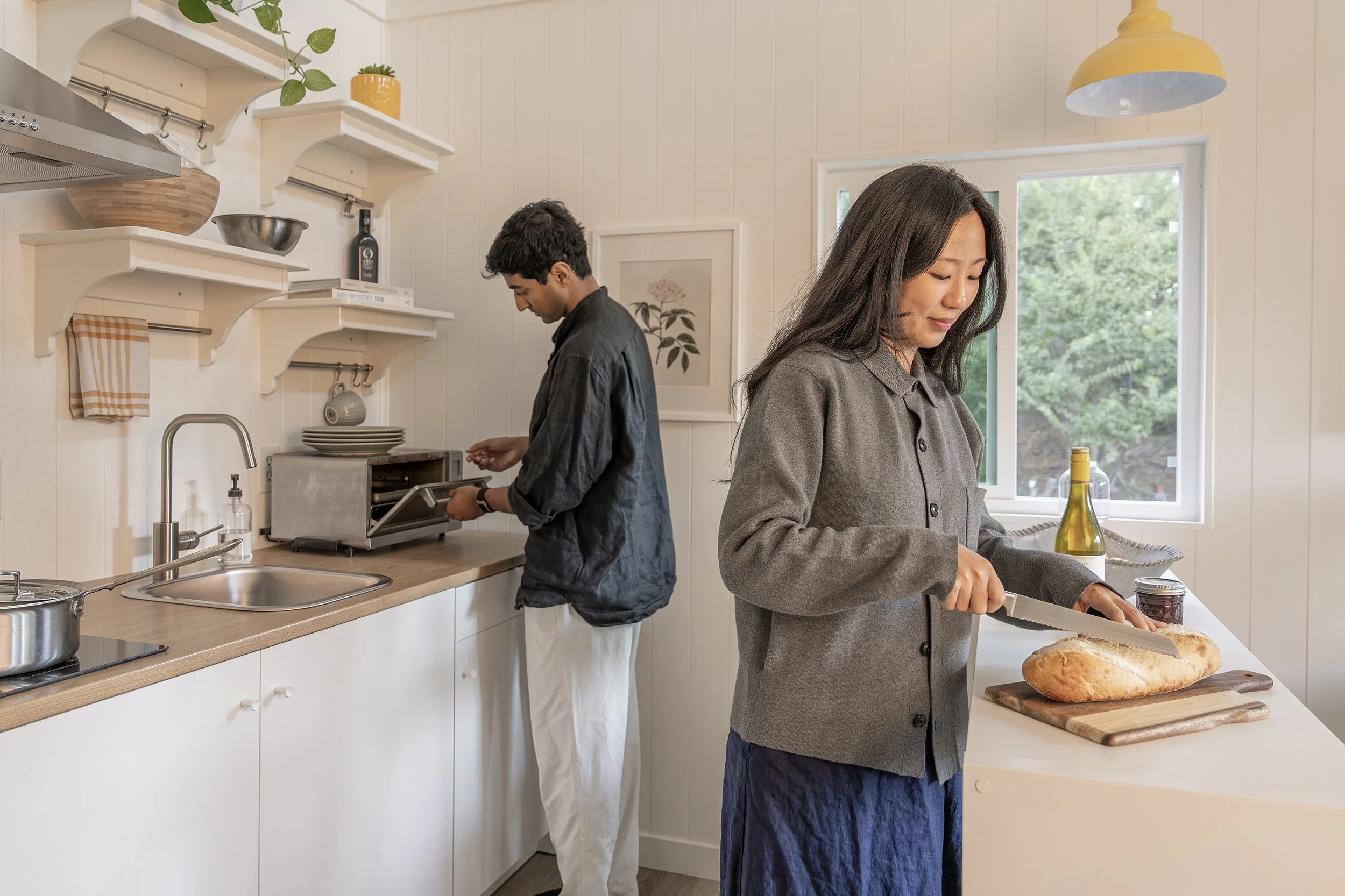 A woman slices bread on a kitchen countertop with a loaf of bread in front of her, while a man is toasting bread in a toaster oven in the background.