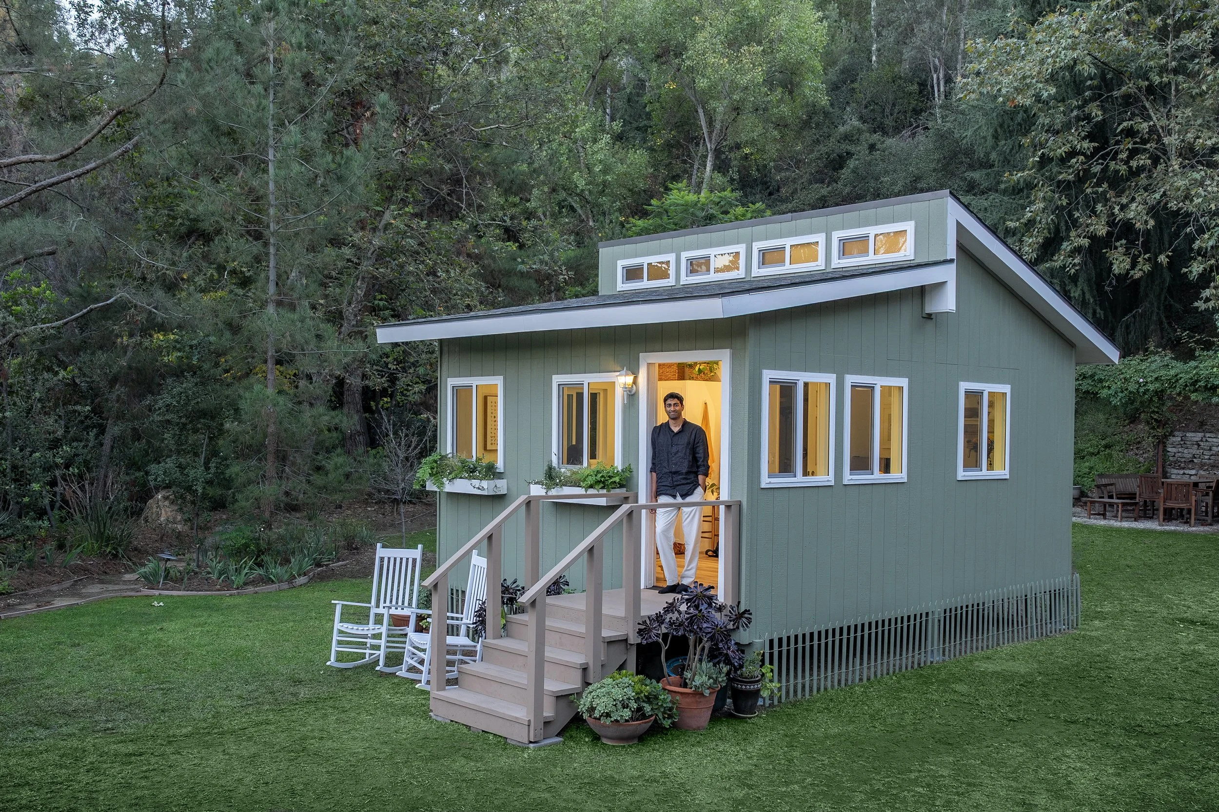 A man stands on the porch of a small green house with a slanted roof, surrounded by lush greenery and a well-kept lawn.