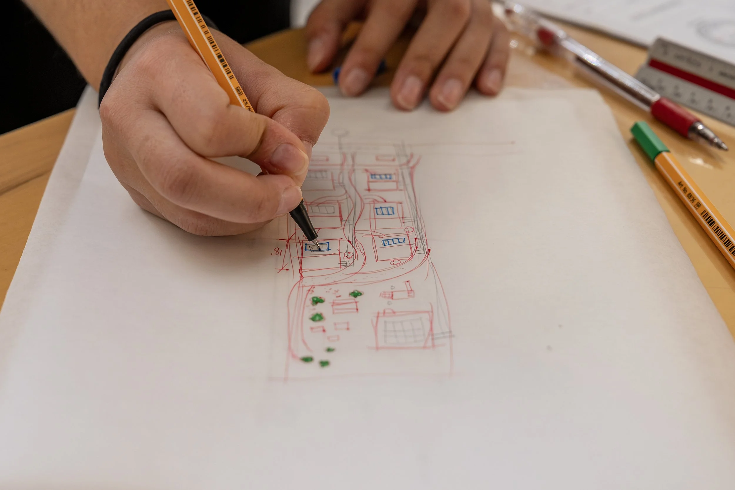 Person drawing a landscape plan with colored pens on white paper, showing houses, landscaping, and a sports court.