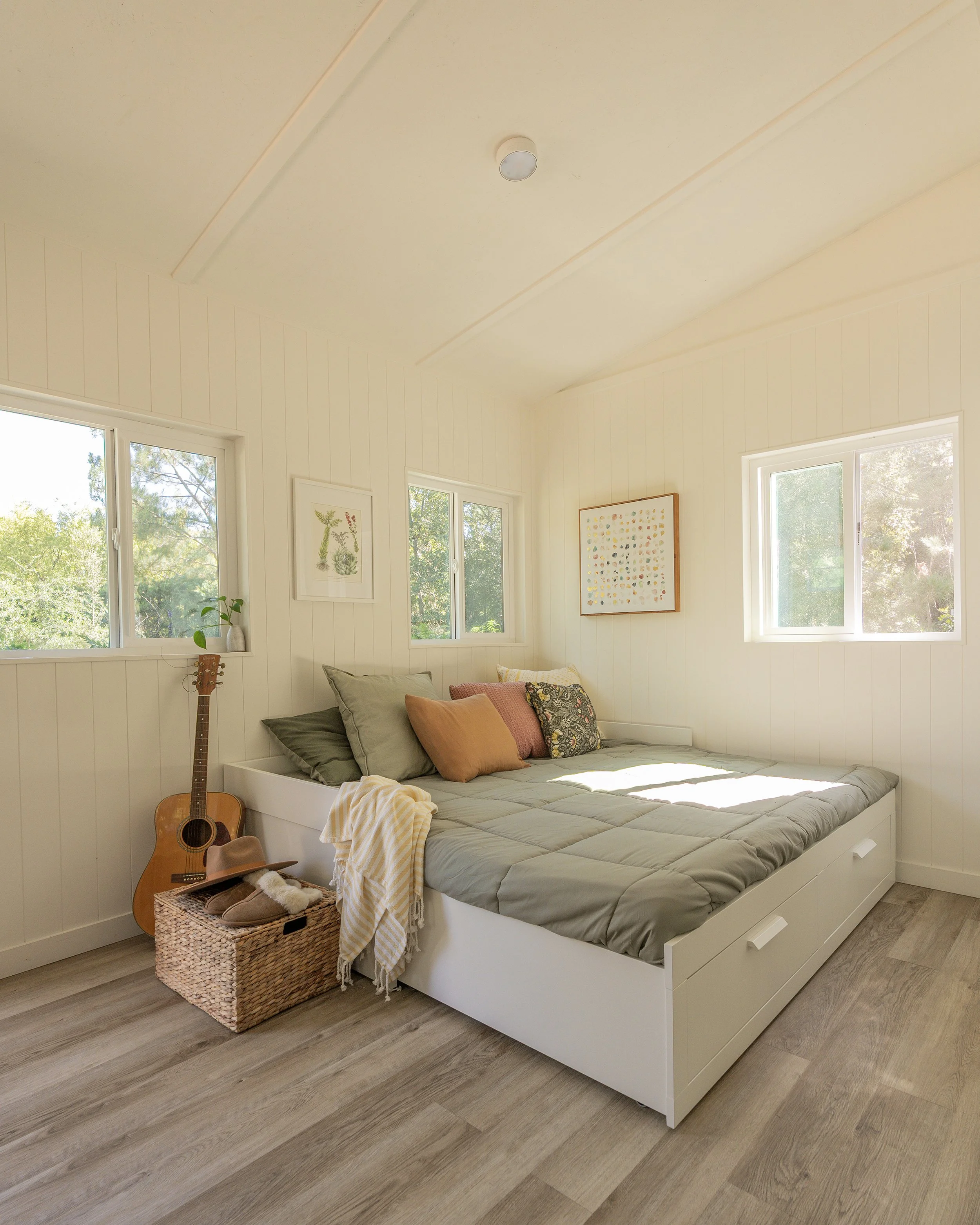 Bright bedroom with white walls, a bed with gray bedding and multiple pillows, three windows letting in natural light, wall art, a guitar leaning against the wall, a woven basket with a hat, a pair of slippers, and a hat on top of the basket.