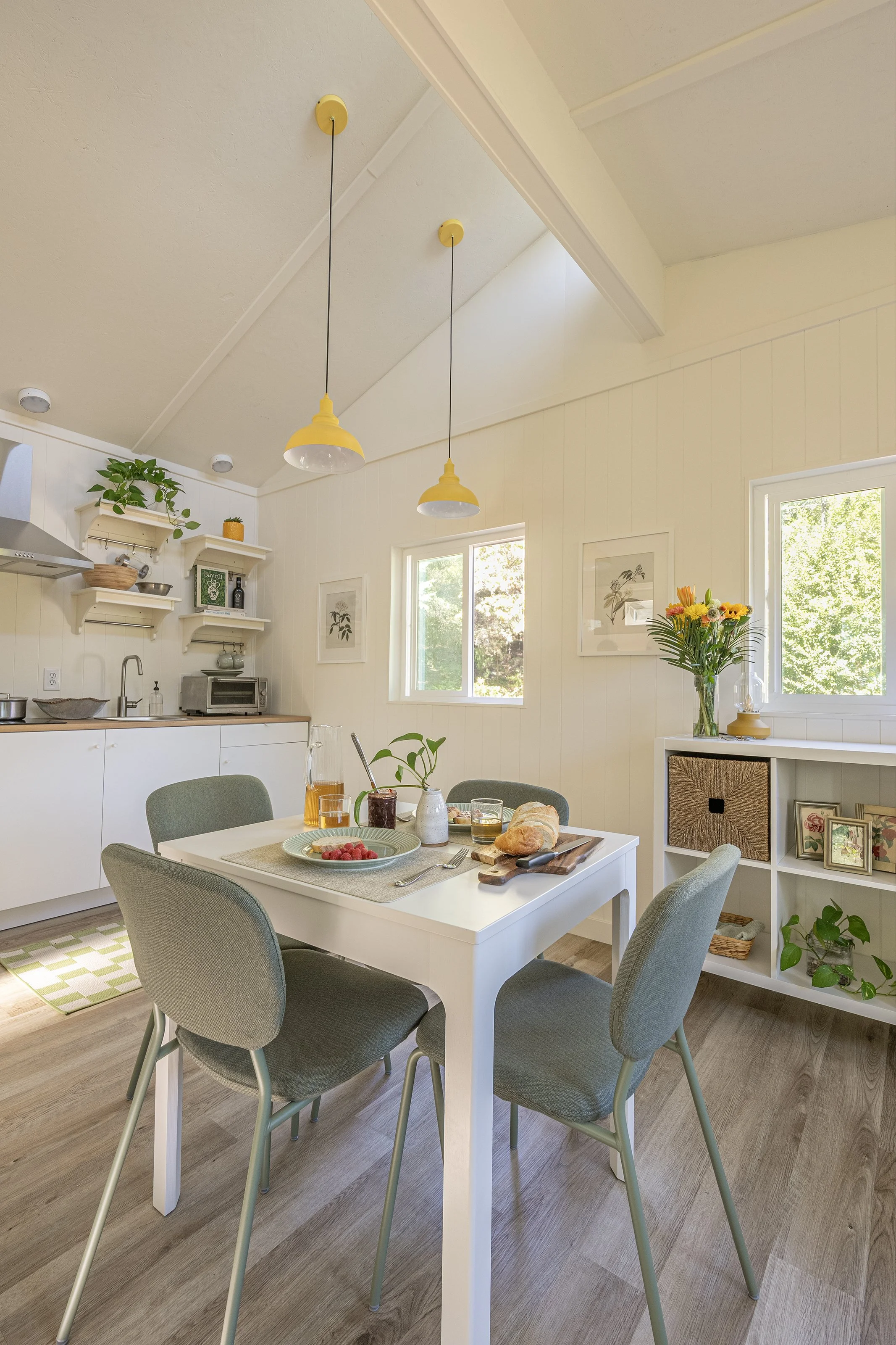 Bright dining area with a white table set for breakfast, surrounded by four green chairs, in a room with wood flooring, light walls, and large windows letting in sunlight, decorated with plants, pictures, and pendant lights.