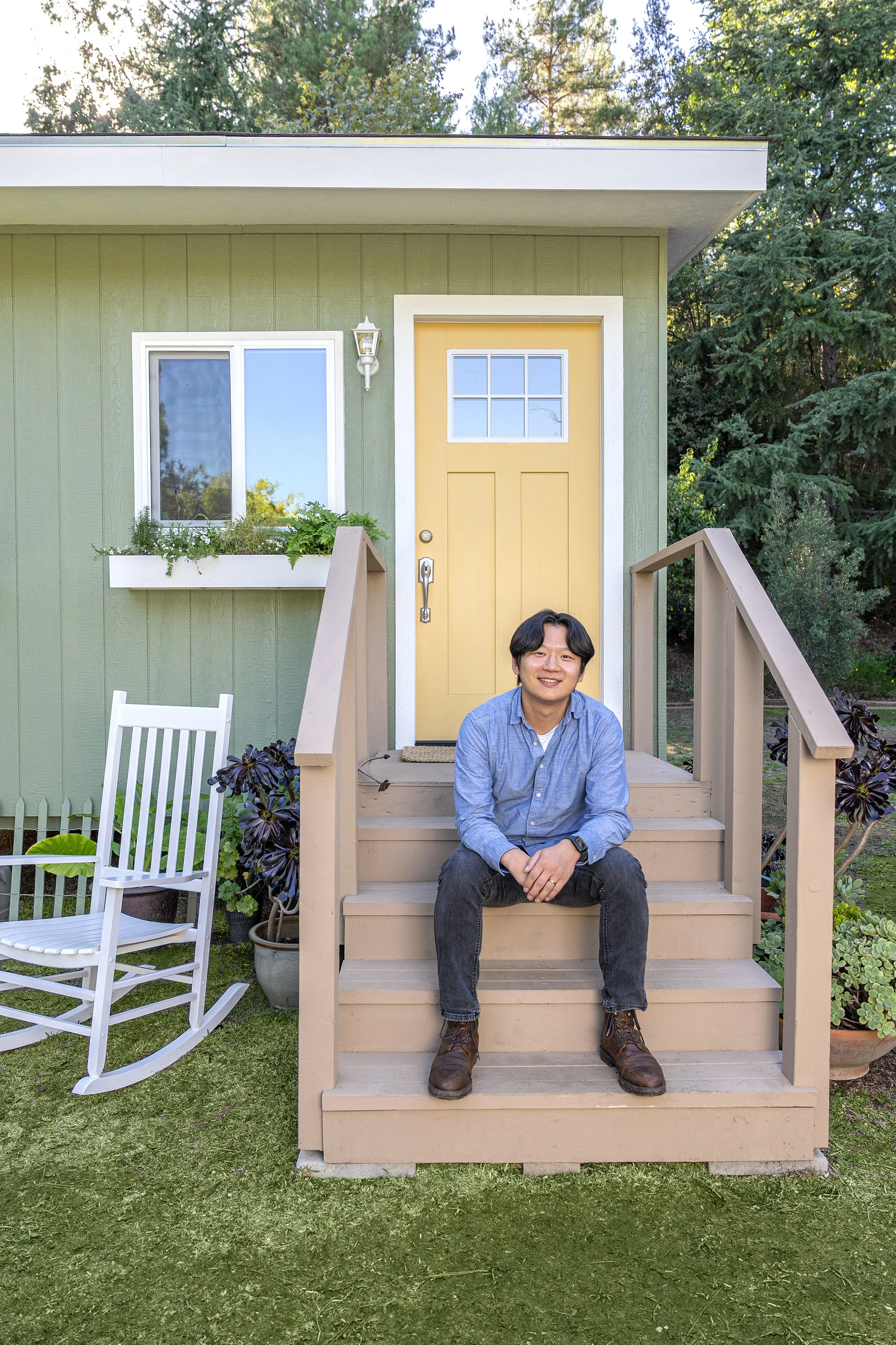 A young man with black hair sitting on the steps of a small front porch in front of a yellow door. The house has green exterior walls, a small white window box with plants, and a white rocking chair on the lawn. The porch has beige stairs and railings, and the background features trees and blue sky.