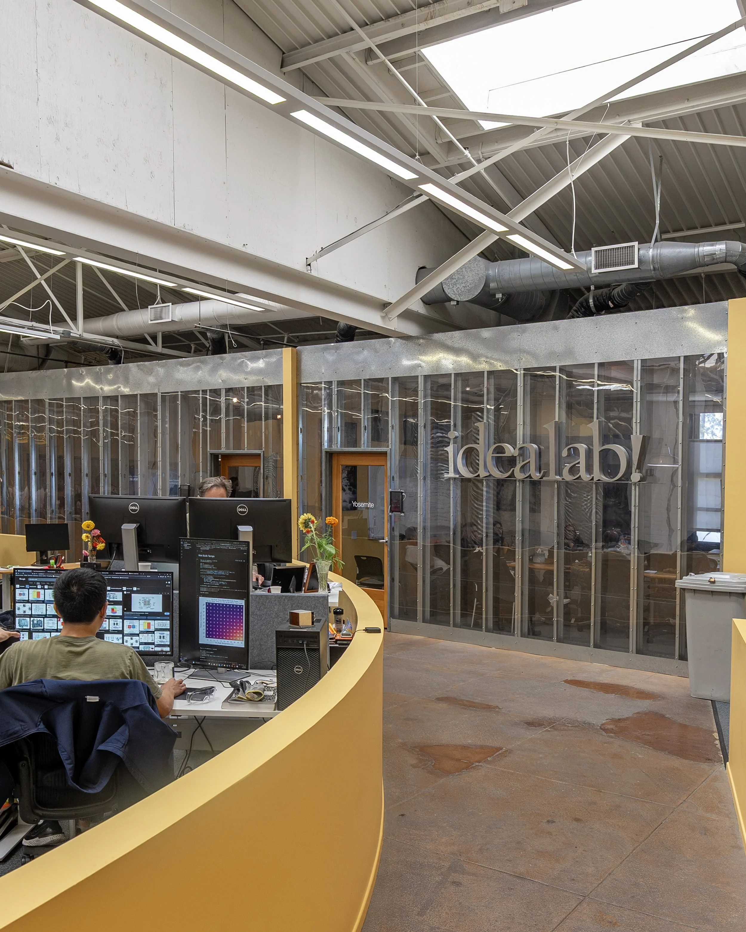 Open office space with multiple computer workstations, a curved yellow desk, and people working at their desks. Office decor includes flowers, and a sign on the back wall reads 'idea ab!'