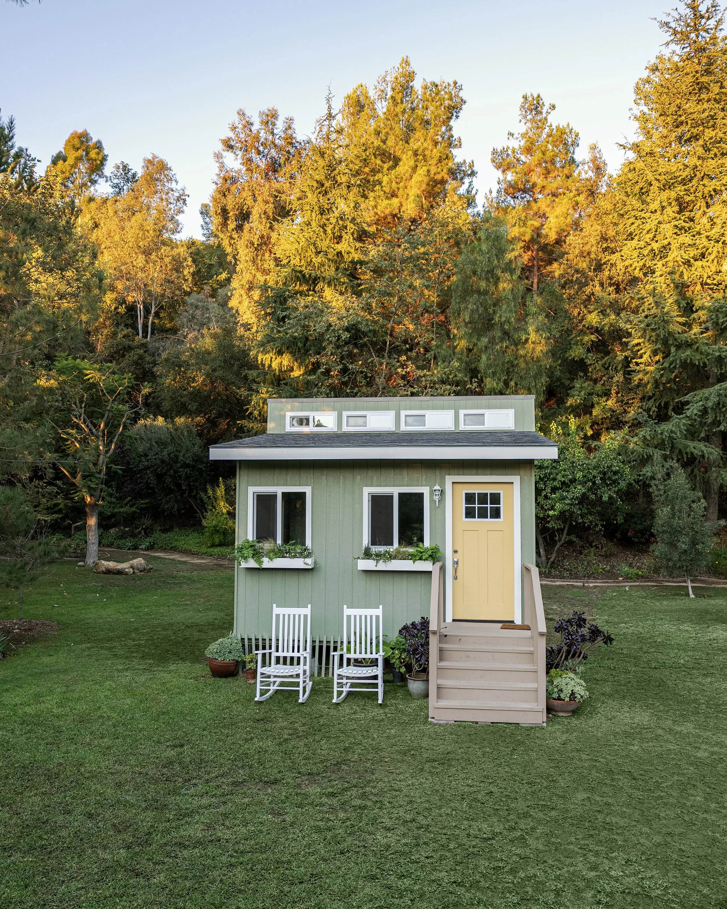Small green house with a yellow door, set on a lawn with potted plants and two white chairs, surrounded by tall trees with autumn-colored leaves.