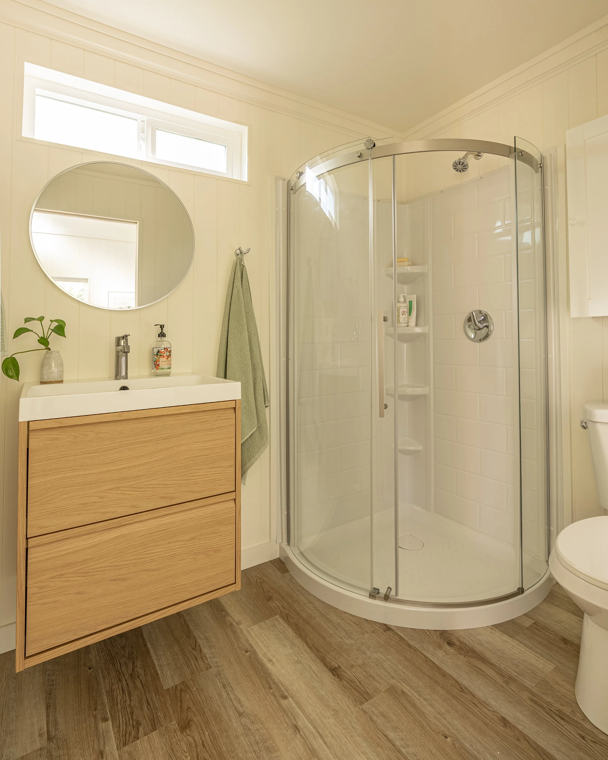 A bathroom with a wooden vanity, a round mirror, a glass corner shower with built-in shelves and white tile walls, a toilet, and a small window near the ceiling letting in natural light.