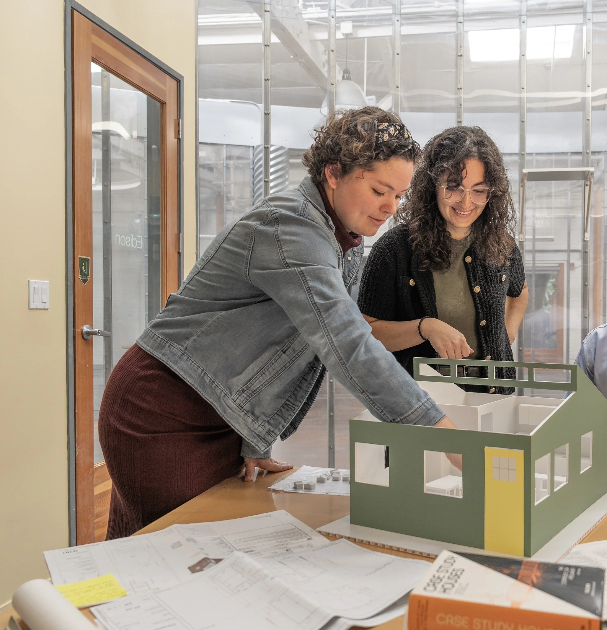 Two women standing over a small model of a green house and smile as they talk.