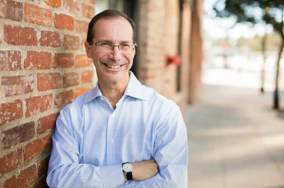 Man with glasses and a blue shirt smiling, standing against a brick wall on a sidewalk.