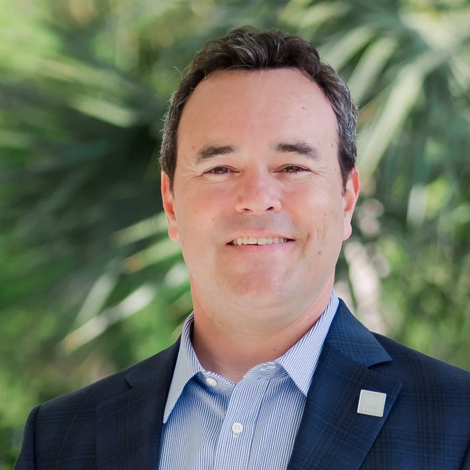 A man with short dark hair smiling, wearing a navy blazer with a pin on the lapel and a light blue collared shirt, standing outdoors with a blurred green foliage background.