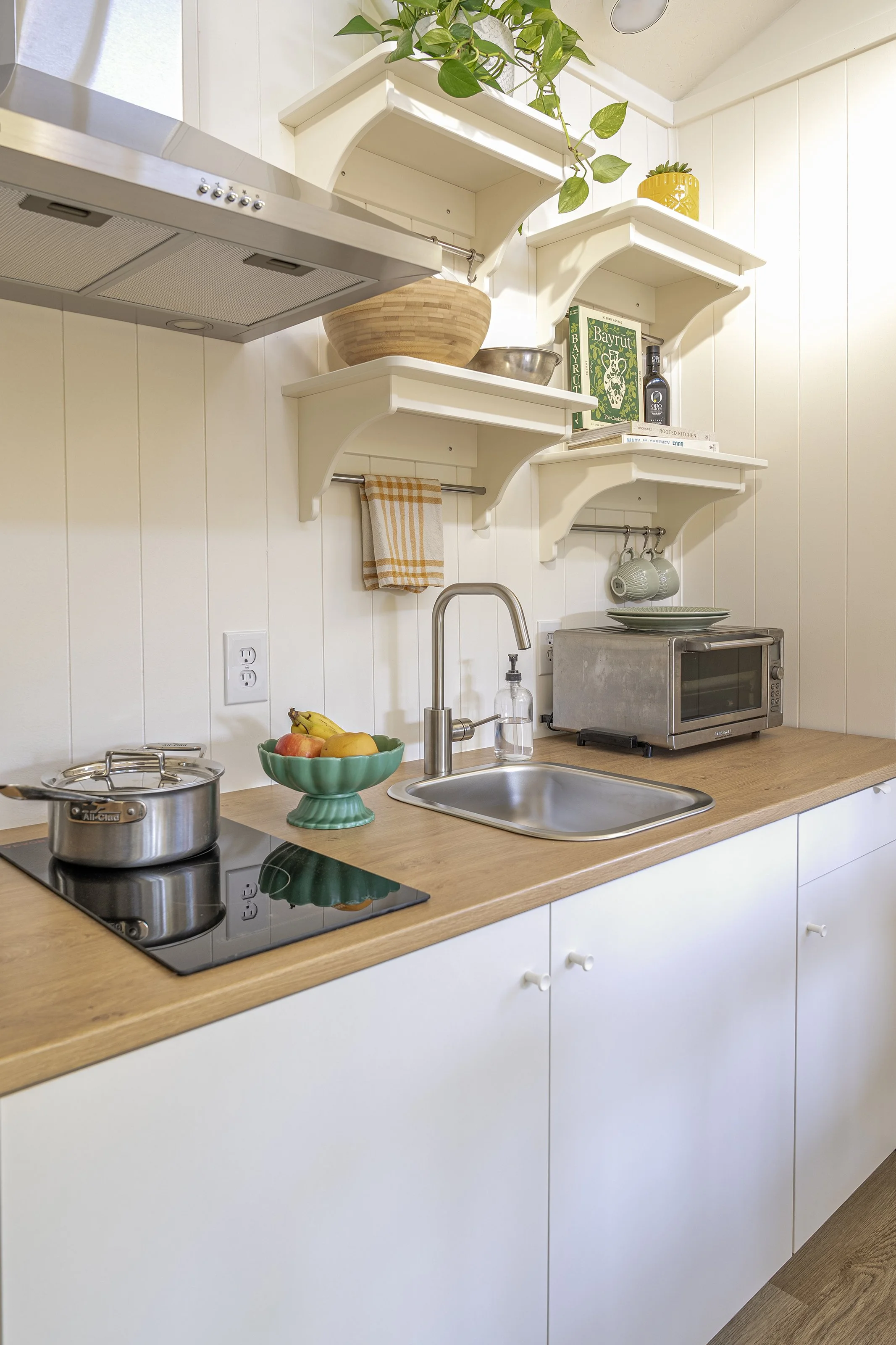 A small, tidy kitchen with white cabinets, a wooden countertop, and open white shelves holding books, bowls, and plants. The counter has a sink, a pot on an induction stove, a fruit bowl, a microwave, and a soap dispenser.