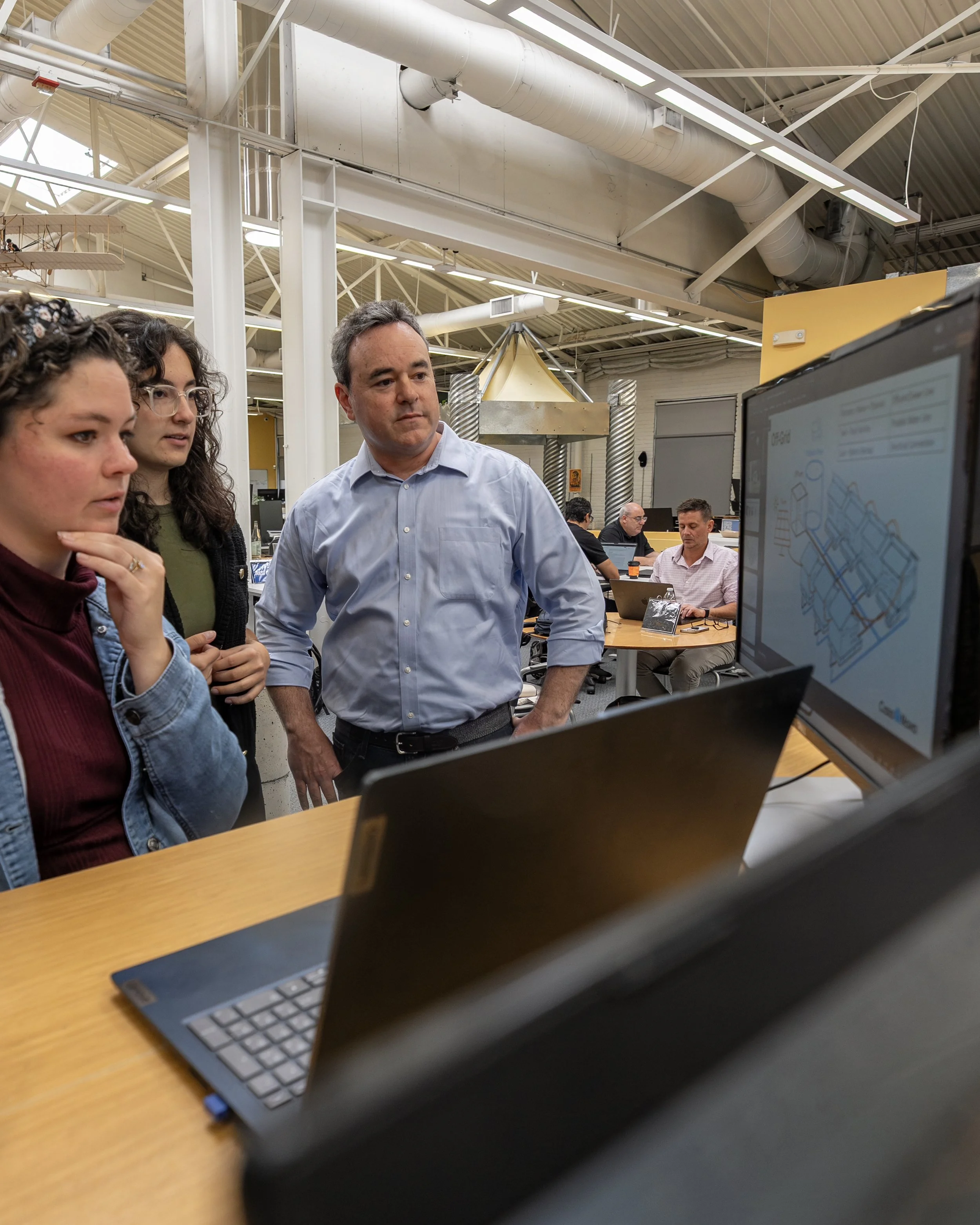 Three people are gathered around a laptop and monitor, looking at a design on the screen, in what appears to be a collaborative work environment or tech workspace.