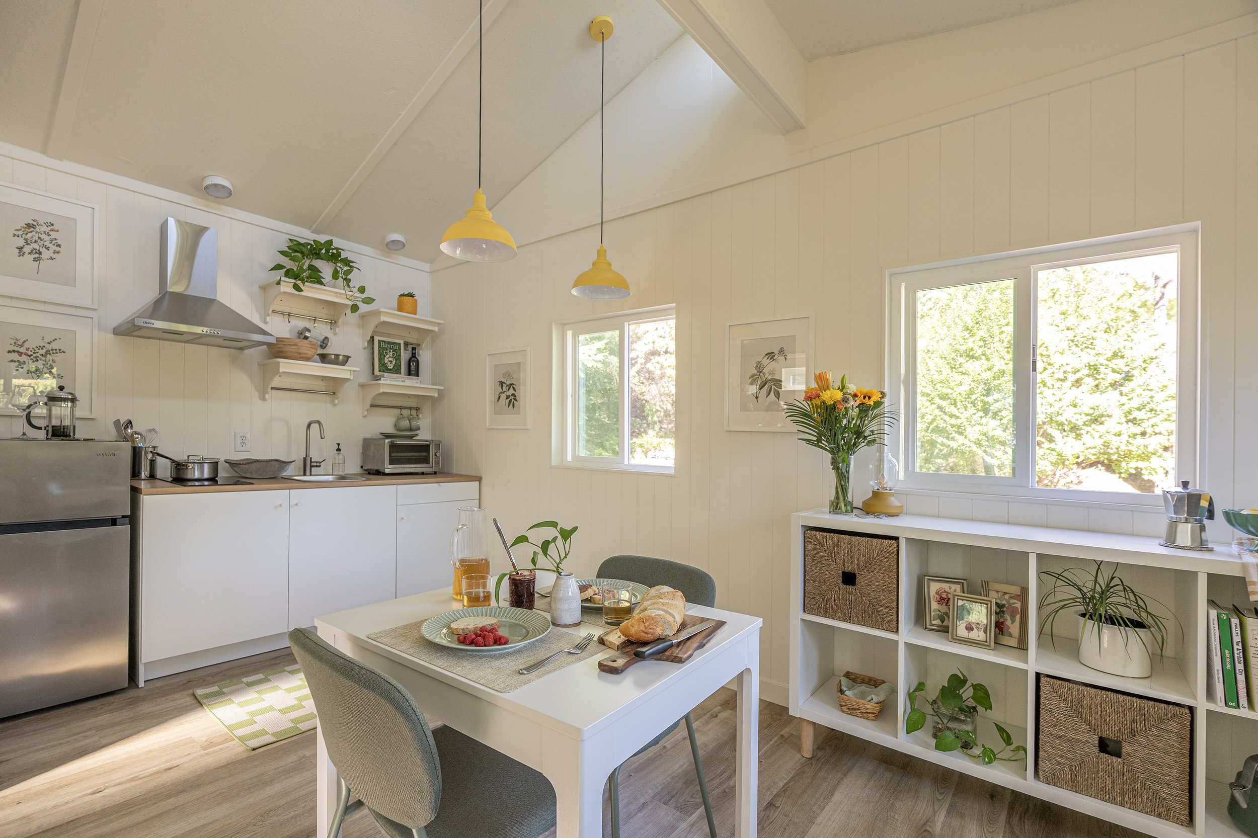 Bright kitchen and dining area with white walls, yellow pendant lights, and a window overlooking greenery. The table is set with a loaf of bread, a bowl of raspberries, and drinks. There are plants and framed artwork on the shelves.