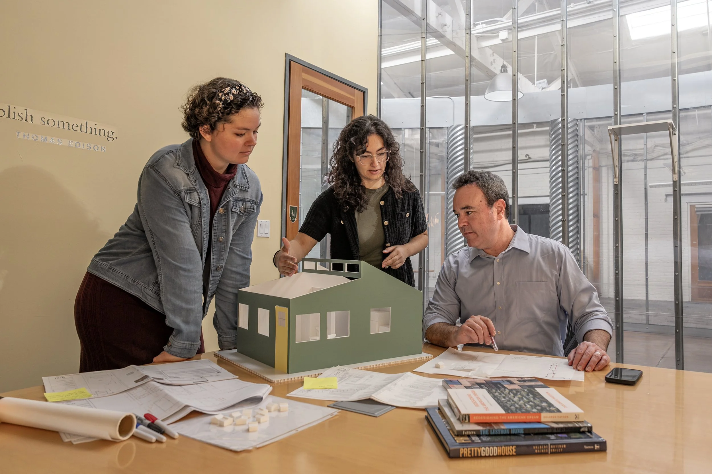 Three people gathered around a table looking at a scale model of a building, with architectural plans and books on the table, in a modern office setting.