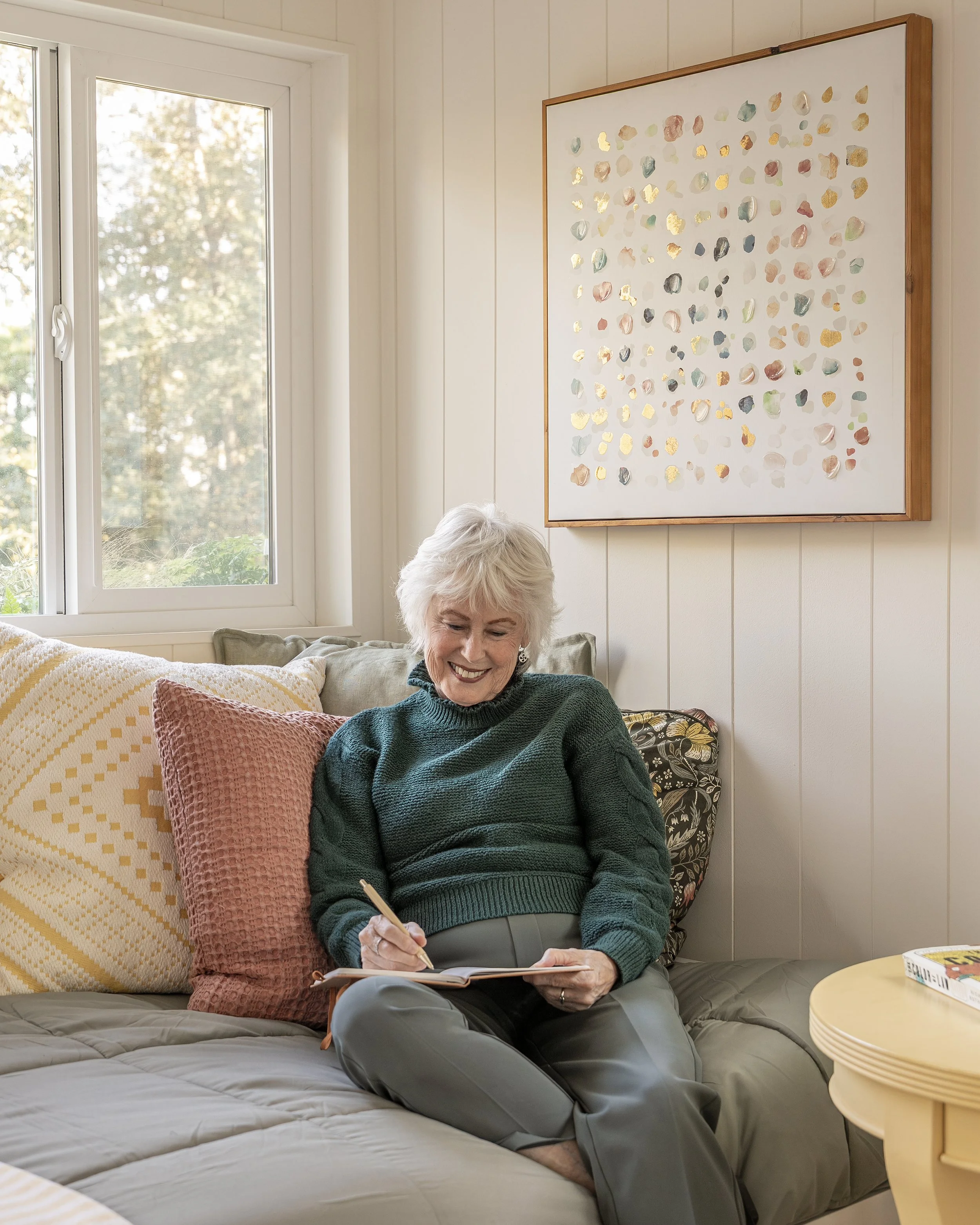 An elderly woman with short white hair sitting on a beige sofa, writing in a notebook, smiling. Behind her is a window with trees outside, and on the wall above her is abstract art with colorful shapes.
