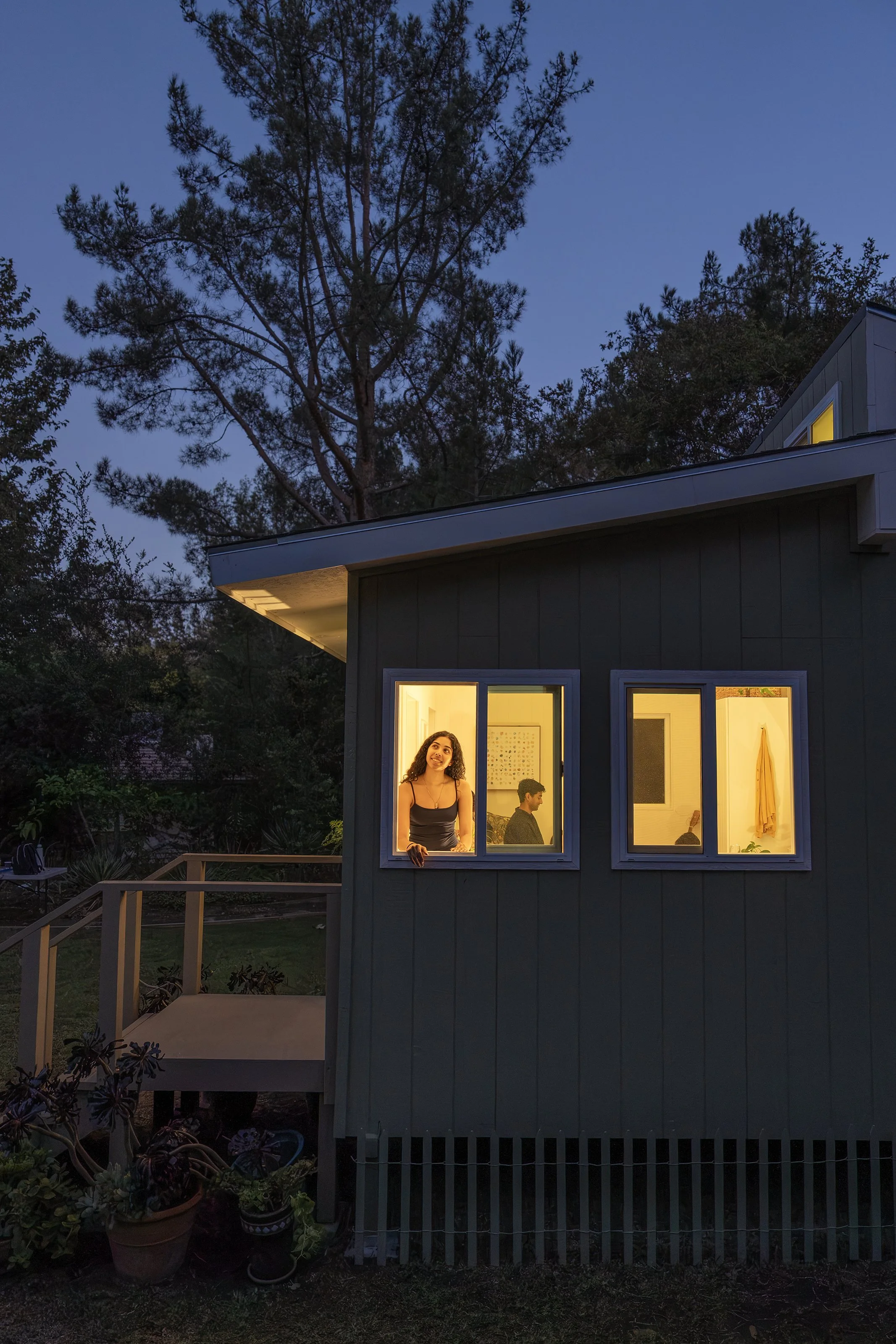 A house with warmly lit windows at dusk, showing a woman leaning out and smiling, with other people inside.