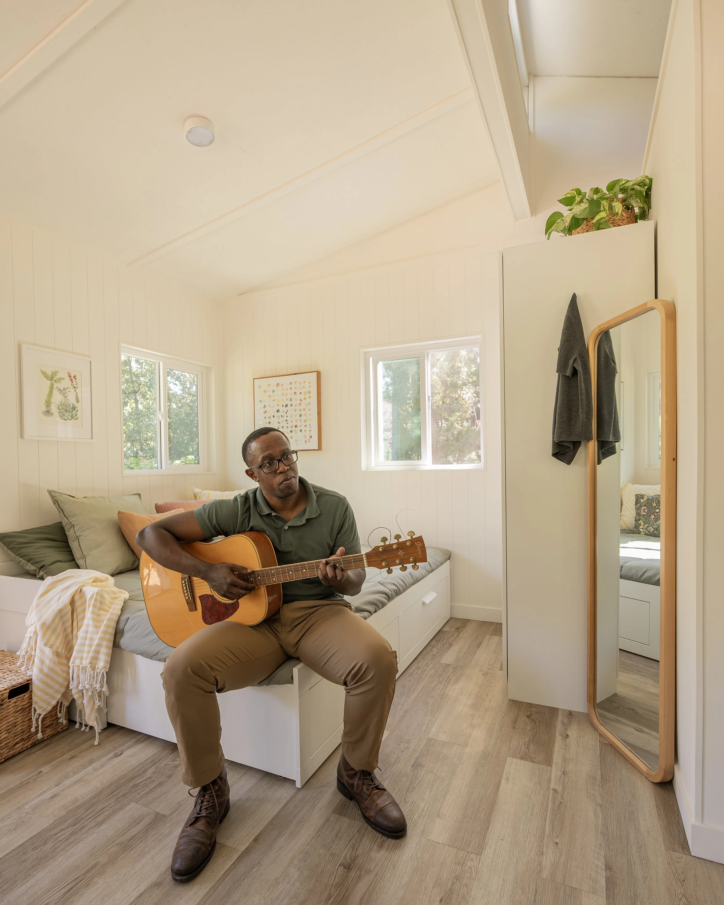 A person sitting on a bed playing an acoustic guitar in a small, bright room with white walls, two windows, a wooden floor, and a full-length mirror.